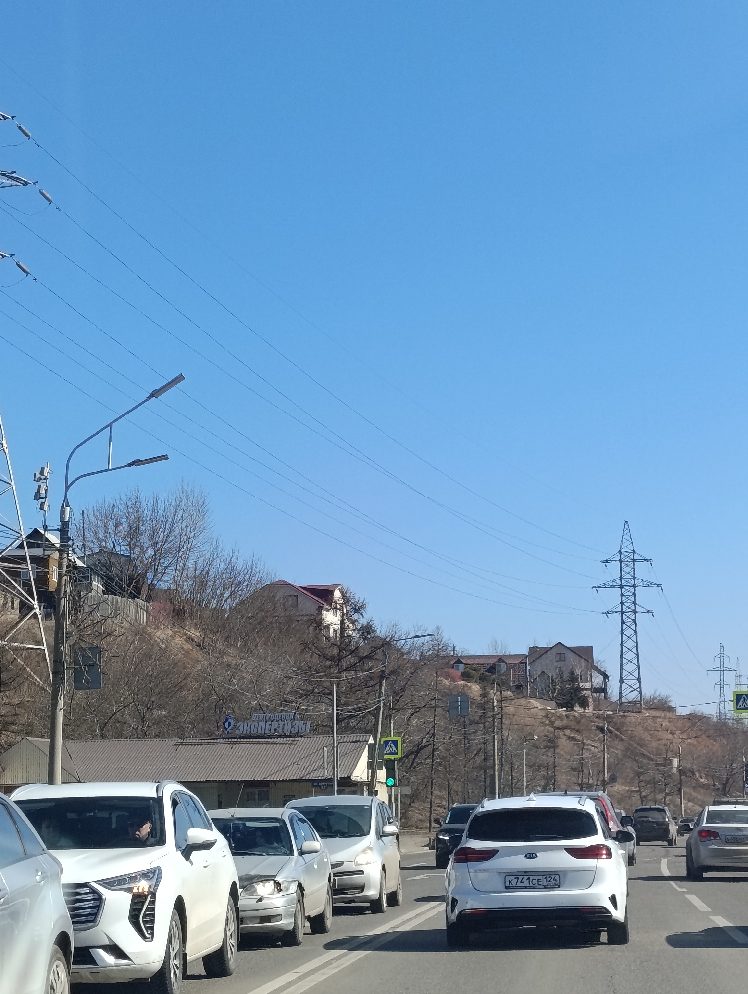 The image depicts a traffic jam on a city street in Krasnoyarsk, Russia. Several cars are visible, including a white Kia with license plate K741CE124. The traffic light is green. Behind the cars, there is a hillside with houses and utility poles. The sign for "Expertise Center" is visible. The sky is clear and blue.