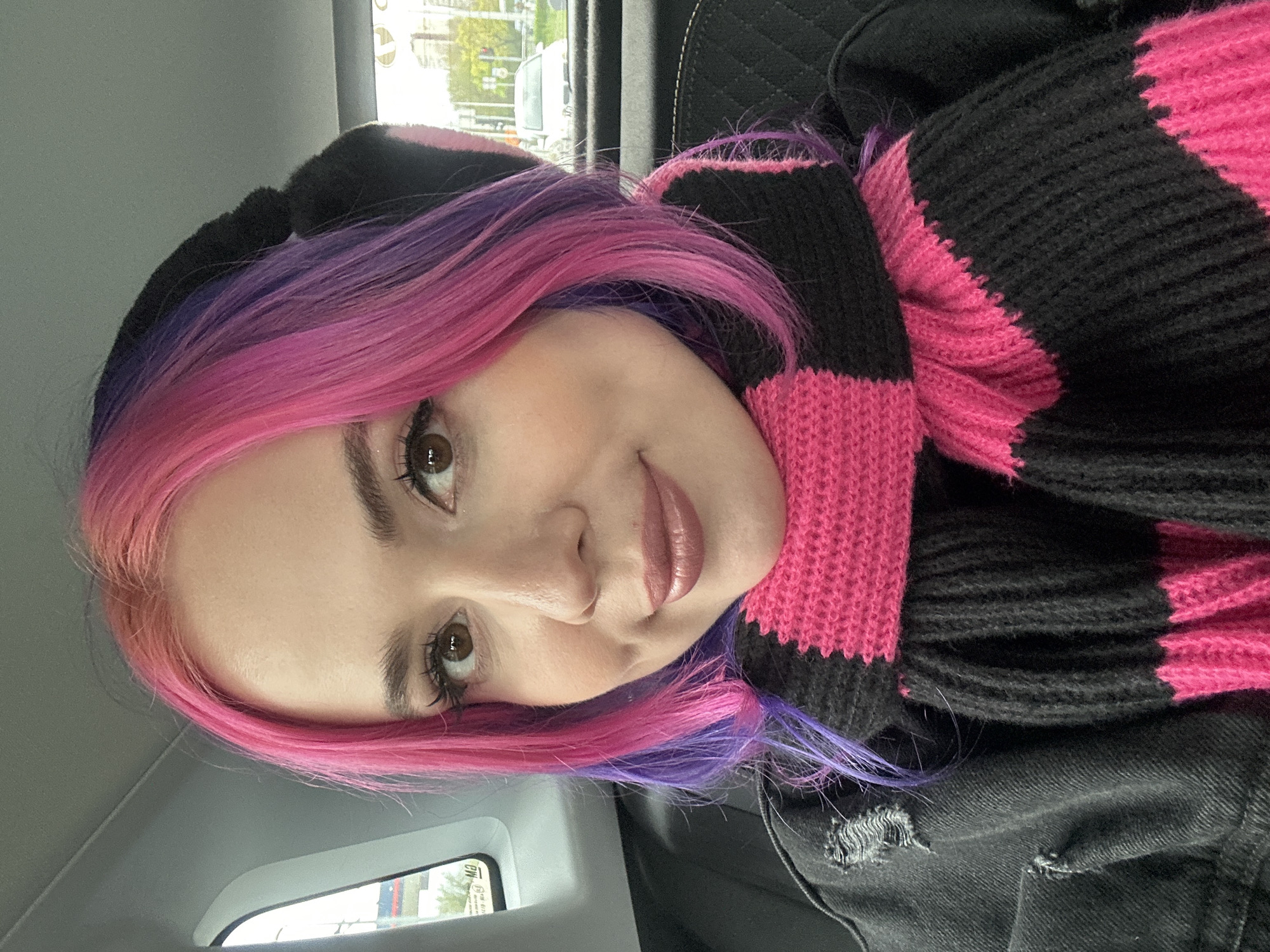 A close-up, angled headshot shows a woman with pink and purple hair. She is wearing a black and pink striped scarf or sweater and a black denim jacket. The setting appears to be inside a car. A window shows blurred city scenery. Based on the clues, this image was taken in City of Zagreb, Croatia.