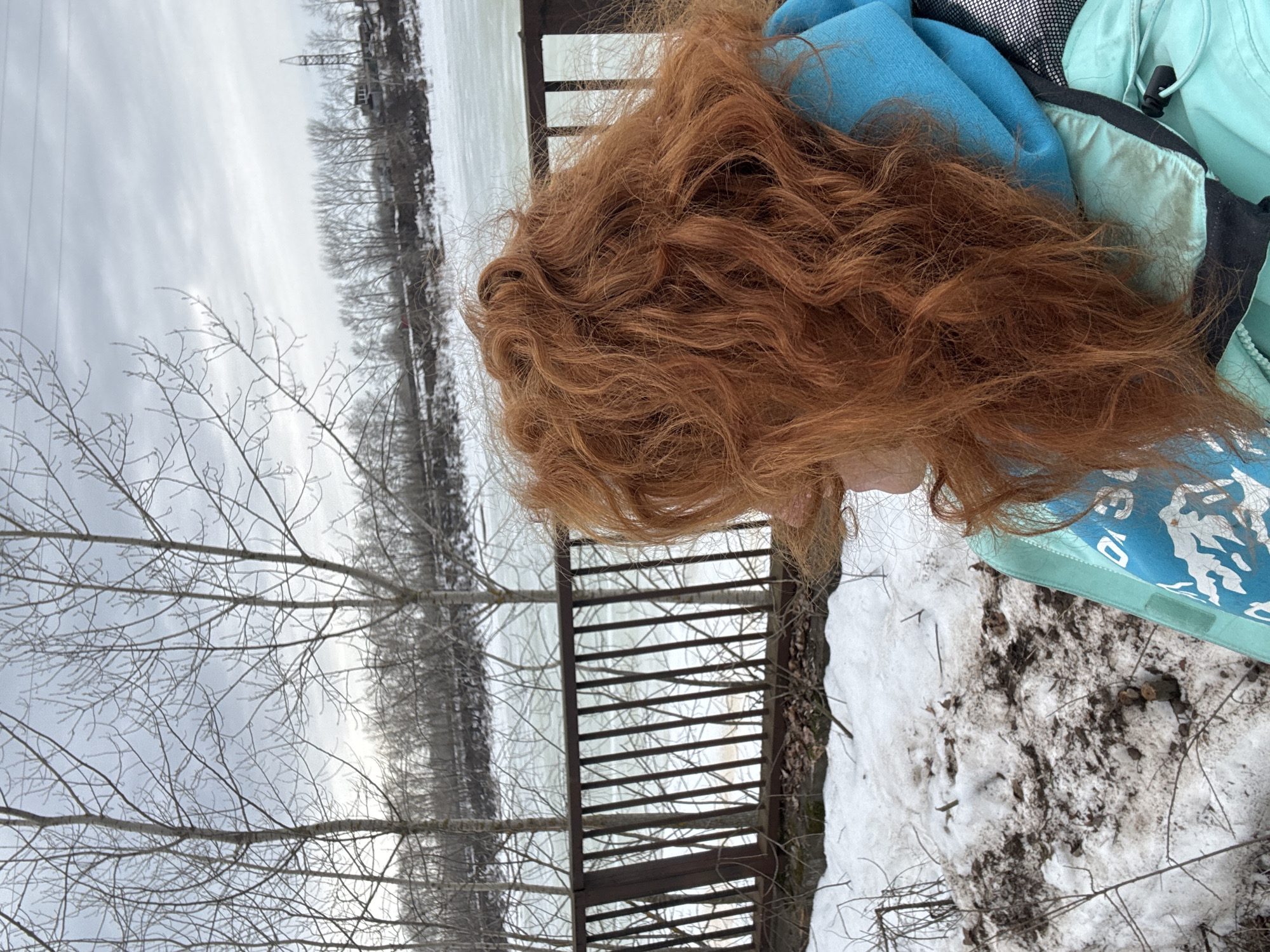 The image presents a partial view of a red-haired person wearing a light blue jacket, likely outdoors in a cold, possibly winter, environment. Snow is present on the ground. A wooden structure resembling a bridge or walkway railing is visible. The background features a body of water, possibly a river or lake, with trees lining the shore. Power lines are also visible in the distance. The location might be in the city of Chaikovsky, Russia.