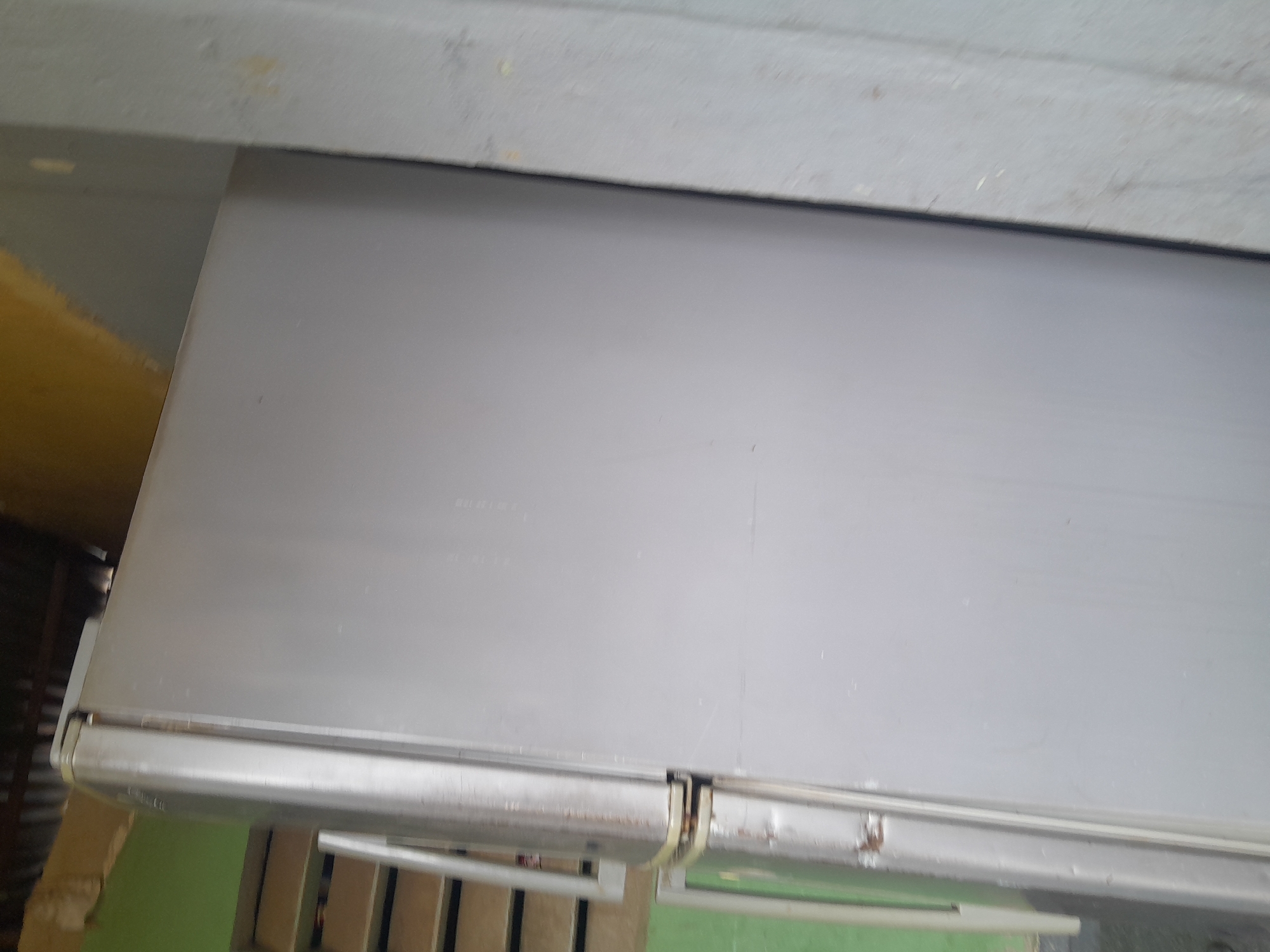 The image shows a light gray, top-freezer refrigerator positioned outdoors. The refrigerator has two doors, with the freezer door appearing to be slightly ajar or at an angle. The exterior of the refrigerator shows some signs of wear and tear, including rust spots, particularly around the door hinges. To the left of the refrigerator, there is a light green wall and a set of stairs with wooden treads. Above the refrigerator, the scene is shadowed, with what appears to be corrugated roofing material visible. To the right of the refrigerator, a light gray, textured surface, possibly a wall or another appliance, runs vertically. The overall environment suggests an outdoor storage area or a less-maintained part of a property in Madalla, Nigeria.