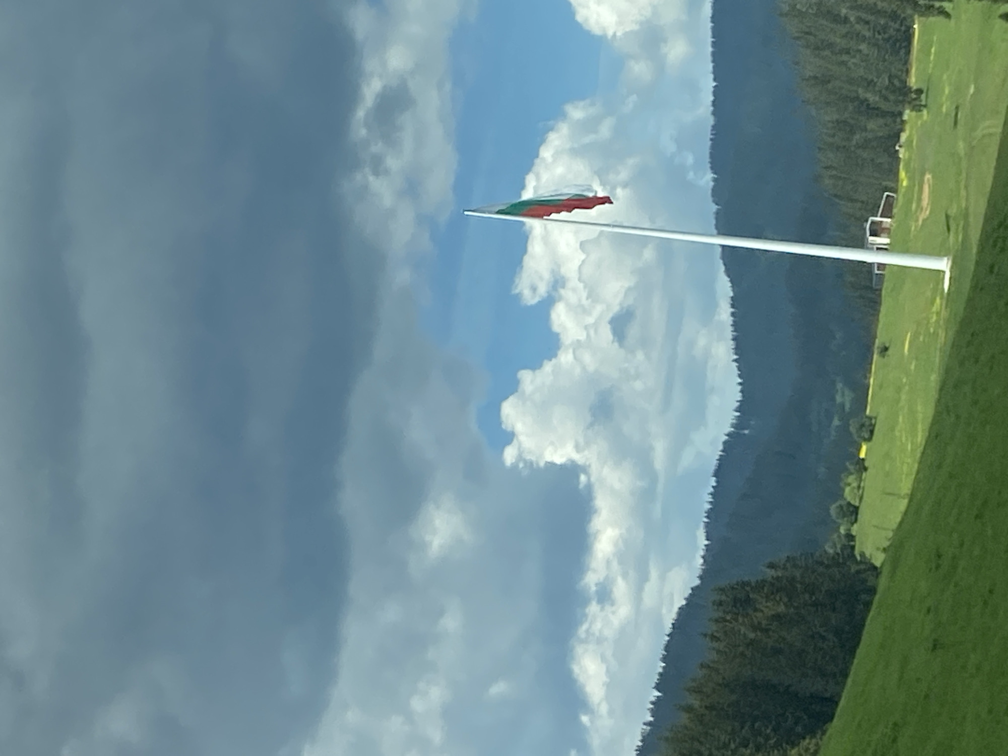 This image captures a vast outdoor landscape under a bright, partly cloudy sky. The scene is dominated by natural elements: a large expanse of green grass in the foreground on the right, rising up to meet a dense, dark green forested mountain range that stretches across the background.
A tall, slender white flagpole stands prominently in the middle-right of the frame, extending vertically through the image. From its top, the flag of Bulgaria is unfurled and gently waving in the breeze. The flag displays three horizontal stripes: white at the top, green in the middle, and red at the bottom.
The sky is a mix of clear blue patches and soft, white cumulus clouds, with some darker, larger clouds gathering towards the upper left, suggesting dynamic weather. In the distance, below the mountain line and to the right of the flagpole, a light-colored structure with distinct rectangular shapes, possibly a building or part of a larger facility, is visible on the green terrain. The overall impression is one of a peaceful, expansive natural setting on a pleasant day. The flag clearly identifies the location as Bulgaria, aligning with the provided context of Smolyan. There is no visible text in the image.
