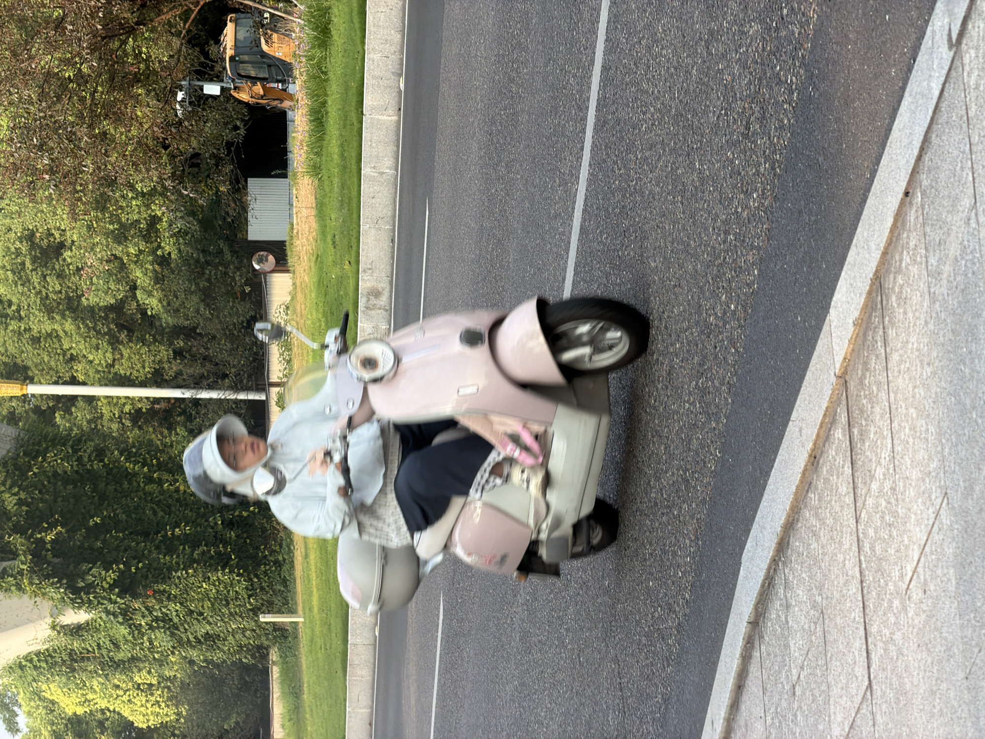 In Jiangjin, China, a person is riding a pink scooter on an asphalt road. The rider is wearing a light-colored jacket, dark pants, and a helmet with a visor. The scooter has a light-colored storage box on the back. The background shows lush green trees, a sidewalk, and a yellow construction vehicle in the distance. The scene appears to be captured during the daytime, with soft lighting.