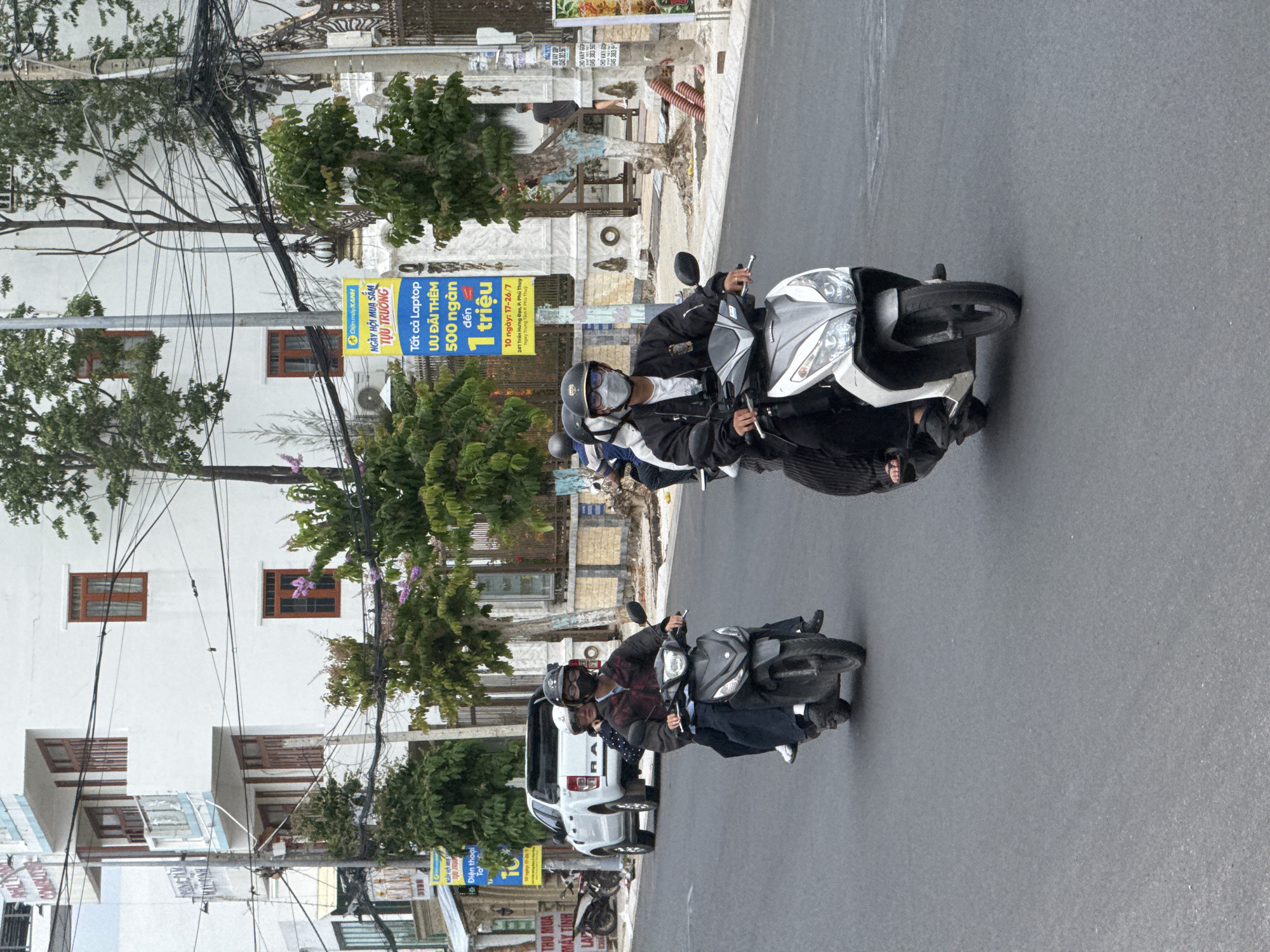 In Phan Thiết, Vietnam, on a paved street, two people ride a scooter. The rider in front wears a dark jacket and a face mask. The passenger behind wears a white helmet and a patterned jacket. The scooter is black and white, and it is in motion. To the left of the scooter, a white pickup truck is parked on the side of the road. A billboard with Vietnamese text and the logo "Điện máy XANH" is visible behind the scooter, advertising a sale on laptops. The background features a white building with several windows and trees. The sky is overcast.