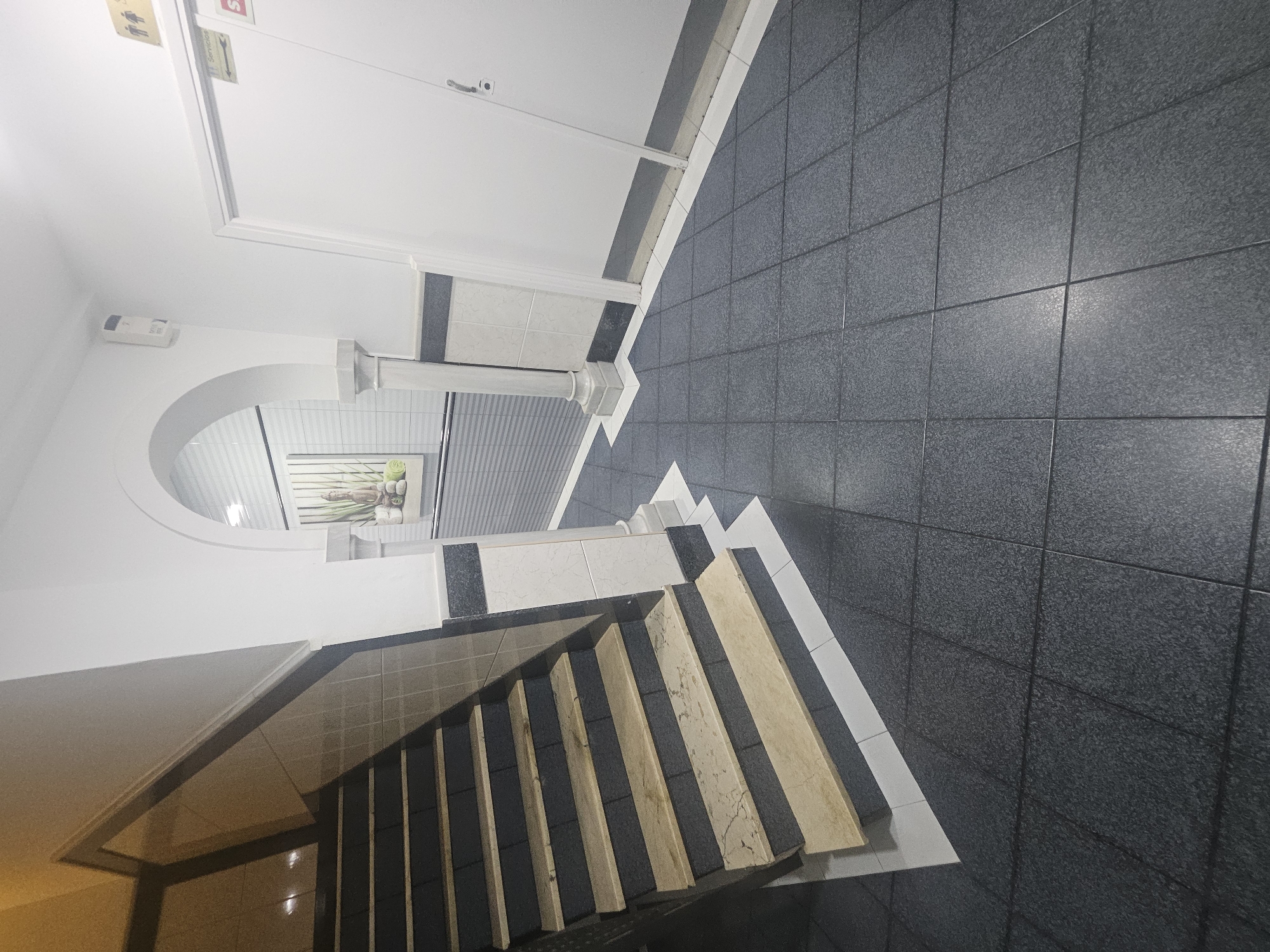 This indoor shot depicts a section of a building interior in Villa del Río, Spain. The scene features a stairway with tile risers leading upward to the right. The stairs are constructed from dark gray tiles and lighter-colored, possibly marble, material for the treads. The stairway is bordered by white trim with black accent tiles. Above the stairs is a rounded archway opening. Behind the archway is a scene depicting a still life with plants. To the right, the floor is covered in dark gray tiles. A white wall is on the left side with a doorway visible. A sign with figures is seen on the door above.