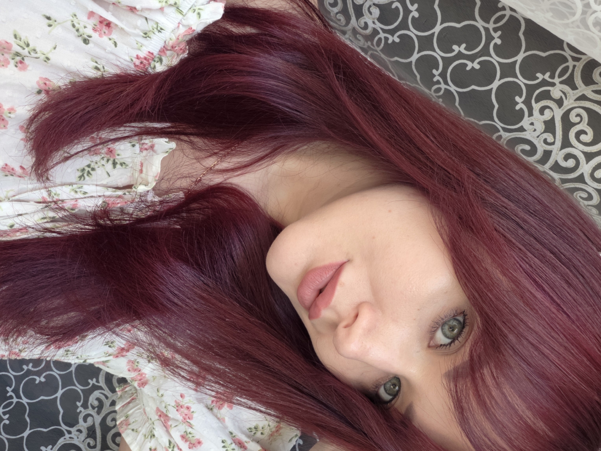 A young woman with long, dark red hair and green eyes is pictured in a close-up shot. She is wearing a white top with a floral pattern in shades of pink and green. Her lips are painted a muted pink color, and she has light makeup on. The background consists of a patterned curtain, with a light-colored section on the left and a darker section with a swirling design on the right. There is no identifiable location in the city of Chaikovsky, Russia.