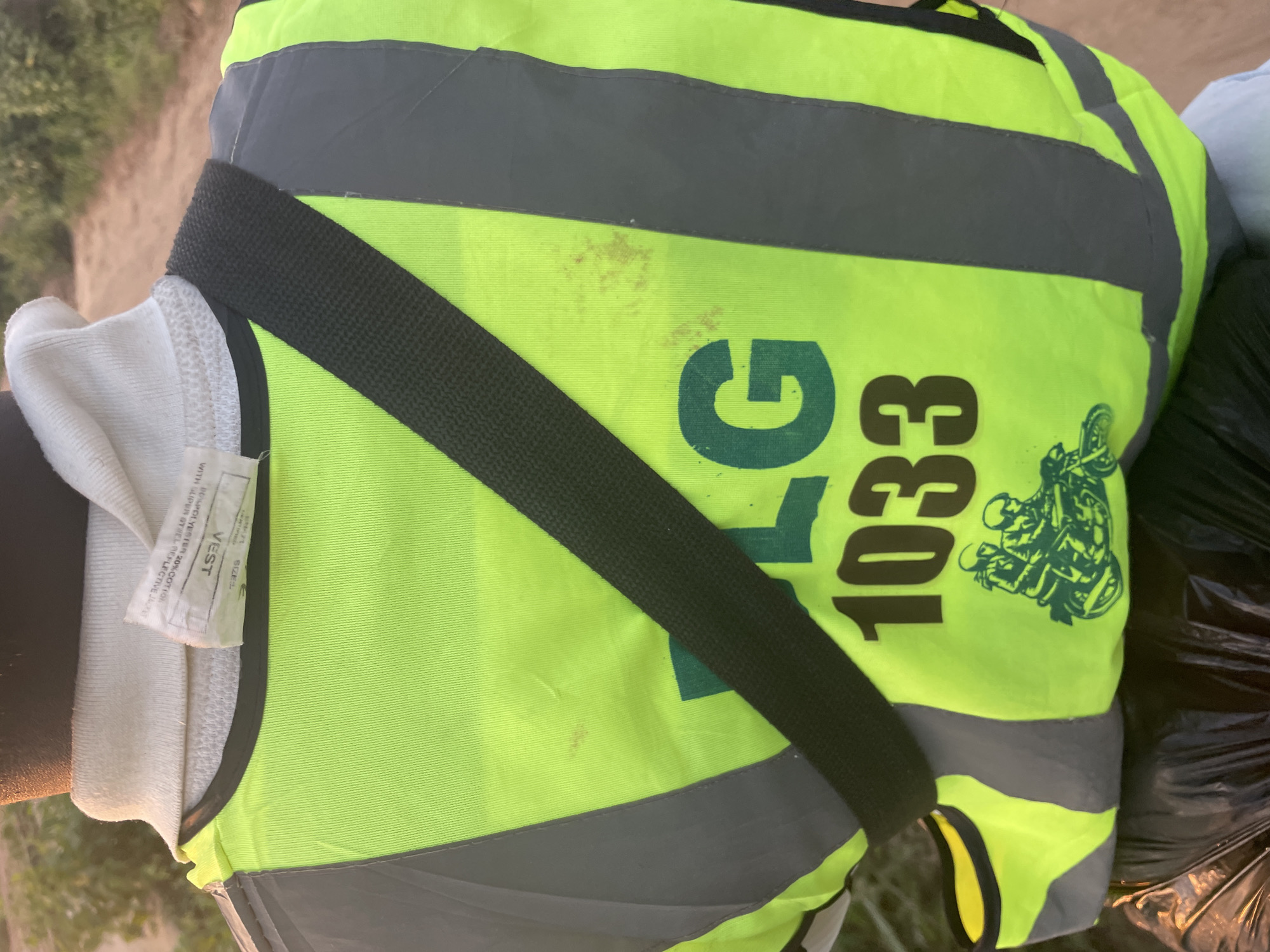 The image shows the back of a person wearing a neon yellow vest with gray reflective stripes. A black strap runs diagonally across the vest. There is green lettering on the vest that reads "PLG 1033" and a green graphic of two people on a motorcycle. A label is attached to the collar of the vest. The background appears to be outdoors with vegetation and a dirt path. This image was taken in Iyafin, Nigeria.