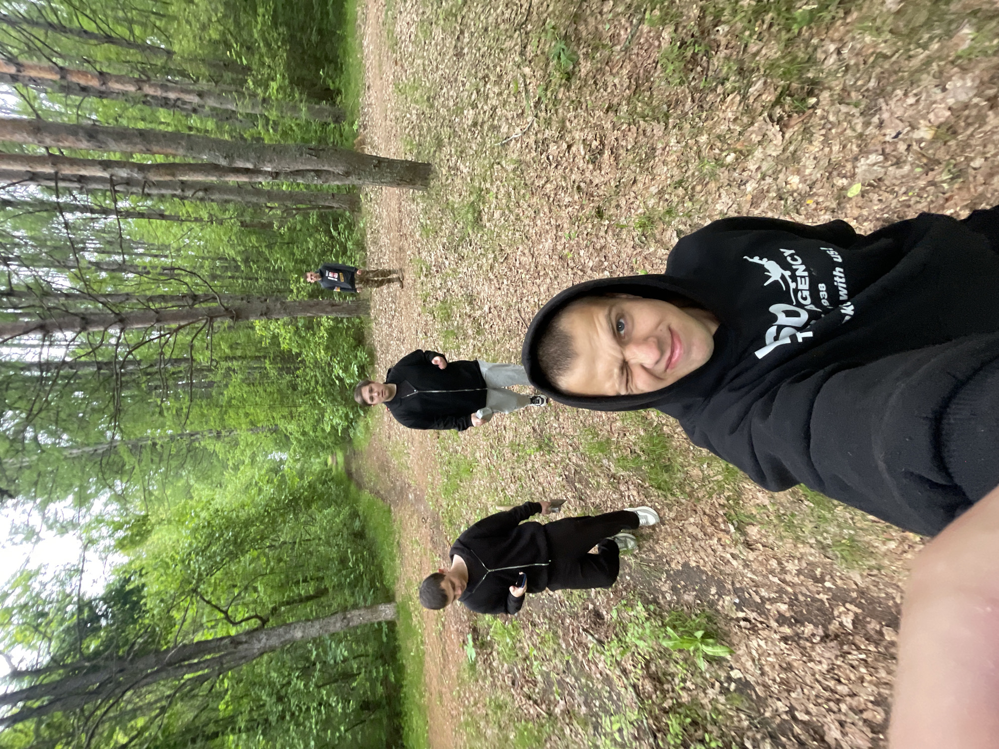 An outdoor selfie captures a group of four individuals on a wooded path in Красные Баки, Russia. The image is taken from a low angle, looking upwards along the path.
In the immediate foreground, a young man with a slight smirk and blue eyes is taking the selfie. He wears a black hooded sweatshirt with the hood up. Visible white text on his hoodie reads "AGENCY 1938 KICK with us!". His right arm is partially visible on the far right.
Behind him, two other young men are visible on the path. One, closer to the foreground, wears a black full-zip hoodie, light-colored pants, and white sneakers, appearing to hold a small object in his hand. Further down the path, another man in a dark top and dark pants also seems to be holding a white object. In the far background, positioned to the left of the path amidst the trees, is a fourth person, possibly a child, wearing a dark top and patterned pants.
The setting is a forest or dense woodland. The ground is covered with a thick layer of dry brown leaves and some patches of green grass. A clear dirt path runs through the center of the scene. Tall, slender tree trunks with light bark line both sides of the path, reaching upwards into a canopy of lush green foliage. The lighting suggests a bright, possibly overcast, daytime. The overall atmosphere is natural and informal, capturing a moment during an outing in the woods.