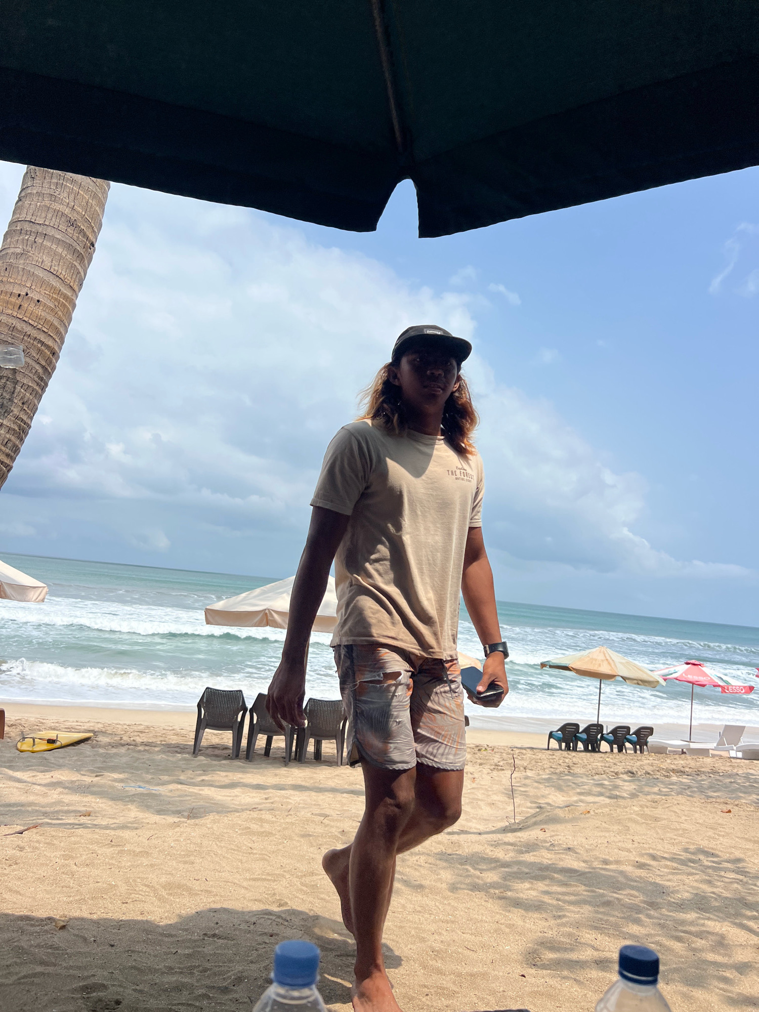 The image depicts a young man with long brown hair, wearing a tan t-shirt, patterned shorts, and a black baseball cap, standing on a sandy beach in Kuta, Indonesia. The man is looking towards the camera and holding a phone in his right hand. The background features the ocean with waves, beach chairs, umbrellas, and a palm tree on the left side. The sky is partly cloudy. A yellow surfboard is visible on the sand in the distance. There are two plastic water bottles in the foreground.