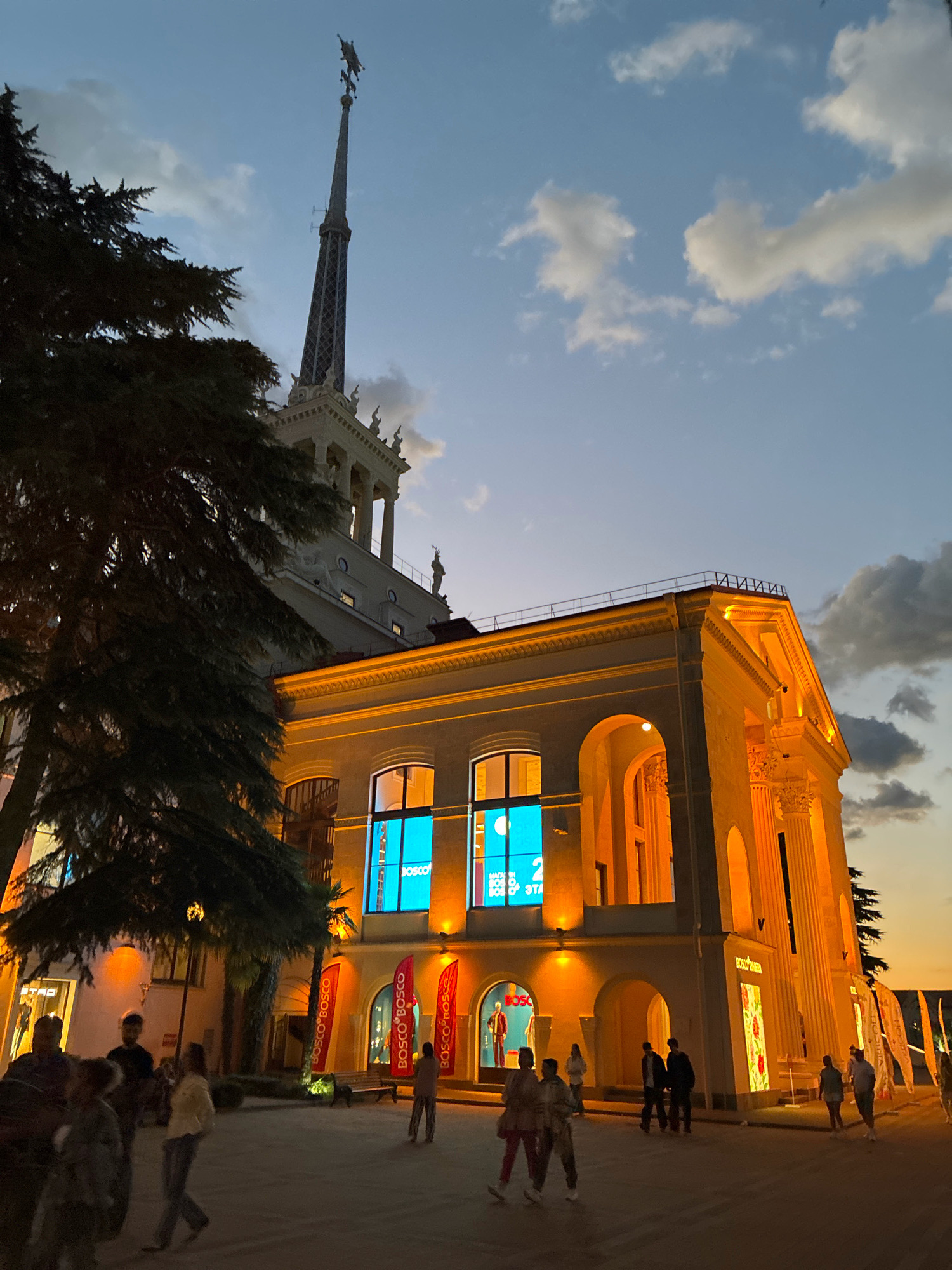 The image depicts the Sochi Sea Port in Sochi, Russia, at dusk. A large, ornate building with classical columns and arches is illuminated by warm, yellow lights. Tall windows on the building display bright blue advertisements. A tall, slender spire topped with a sculpture of a horse and rider rises above the building. A large evergreen tree stands to the left of the building, its dark foliage silhouetted against the twilight sky. Several people are walking on the paved area in front of the building, some in groups and some individually. The sky is a gradient of blue and orange, with scattered white clouds. The scene appears to be a public space, possibly a promenade or plaza, with storefronts and pedestrian activity.