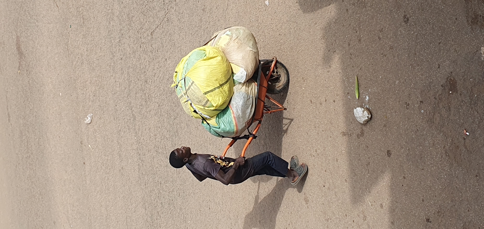 In Bukuru, Nigeria, a young person pushes a wheelbarrow loaded with large bags on a paved surface. The individual, wearing a dark t-shirt and dark trousers, is shown from the side, looking forward. The wheelbarrow, with orange handles and frame, carries several bulky, tied bags, predominantly yellow and green, with some white bags also visible. The paved ground appears to be a road or pathway, with a shadow cast by the person and wheelbarrow. In the foreground, a light-colored rock and a green leaf lie on the ground. The overall scene suggests activity related to transport or collection of goods.