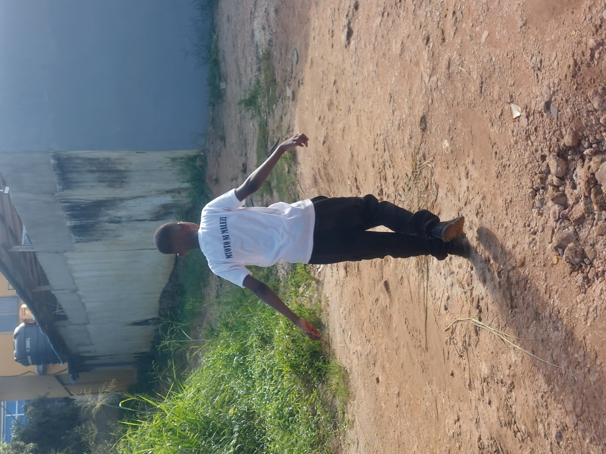 A person is standing on a dirt path. The person is wearing a white shirt with text on the back in Swahili that says, “MTOTO NI MALEZI,” meaning, "A child is brought up." The person is also wearing black pants and brown shoes. The path is lined with grass and weeds on one side and a building on the other. The dirt path has rocks on it. A water tank is in the background. The location appears to be in a low-income area of Dar es-Salaam, Tanzania.