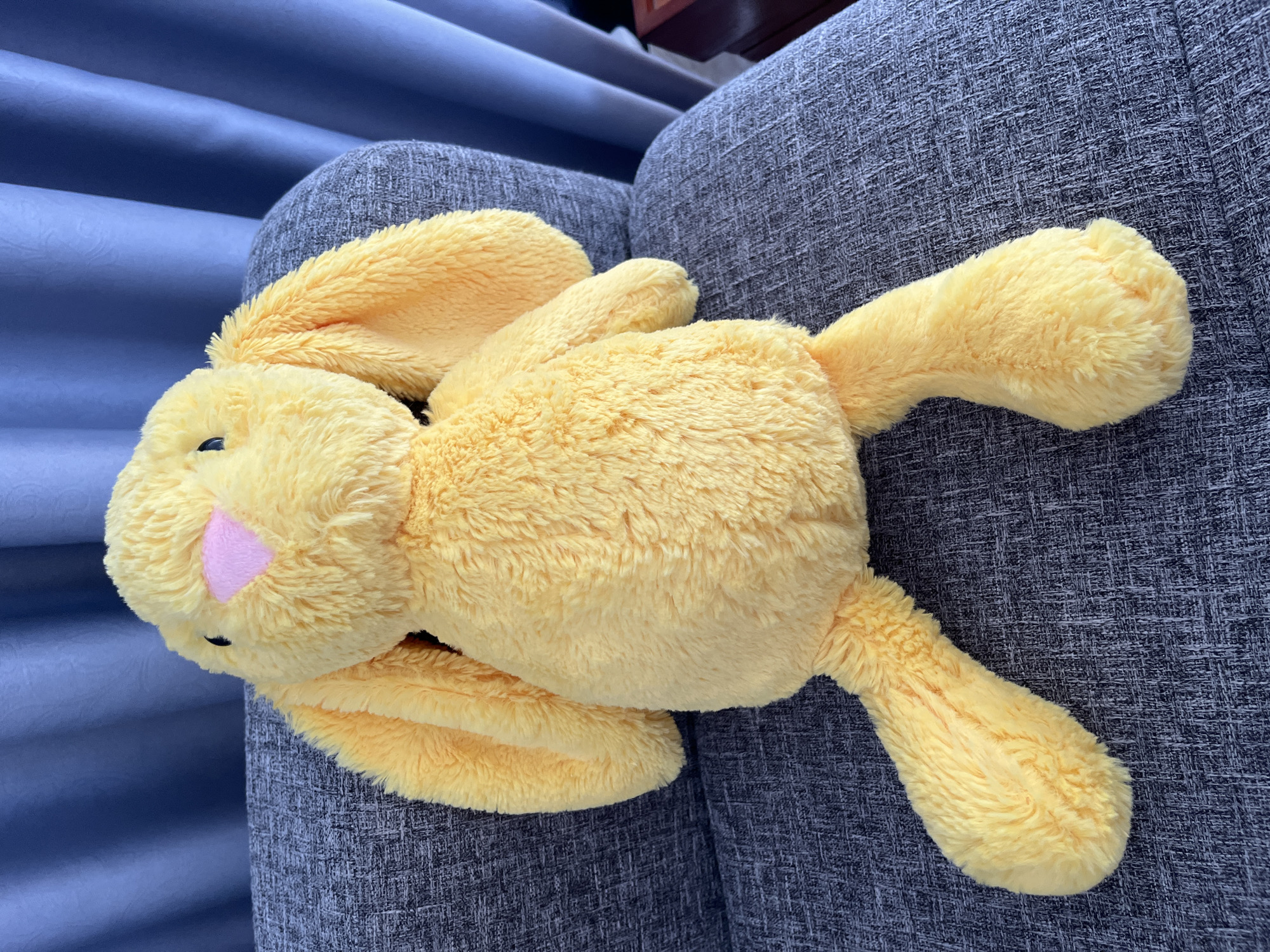 A yellow stuffed bunny lies on a grey upholstered sofa. The bunny has a pink nose and floppy ears. The sofa is in front of a blue curtain. No people are present in the image. There is no indication that the image was taken in Bac Nha Trang Ward, Vietnam.