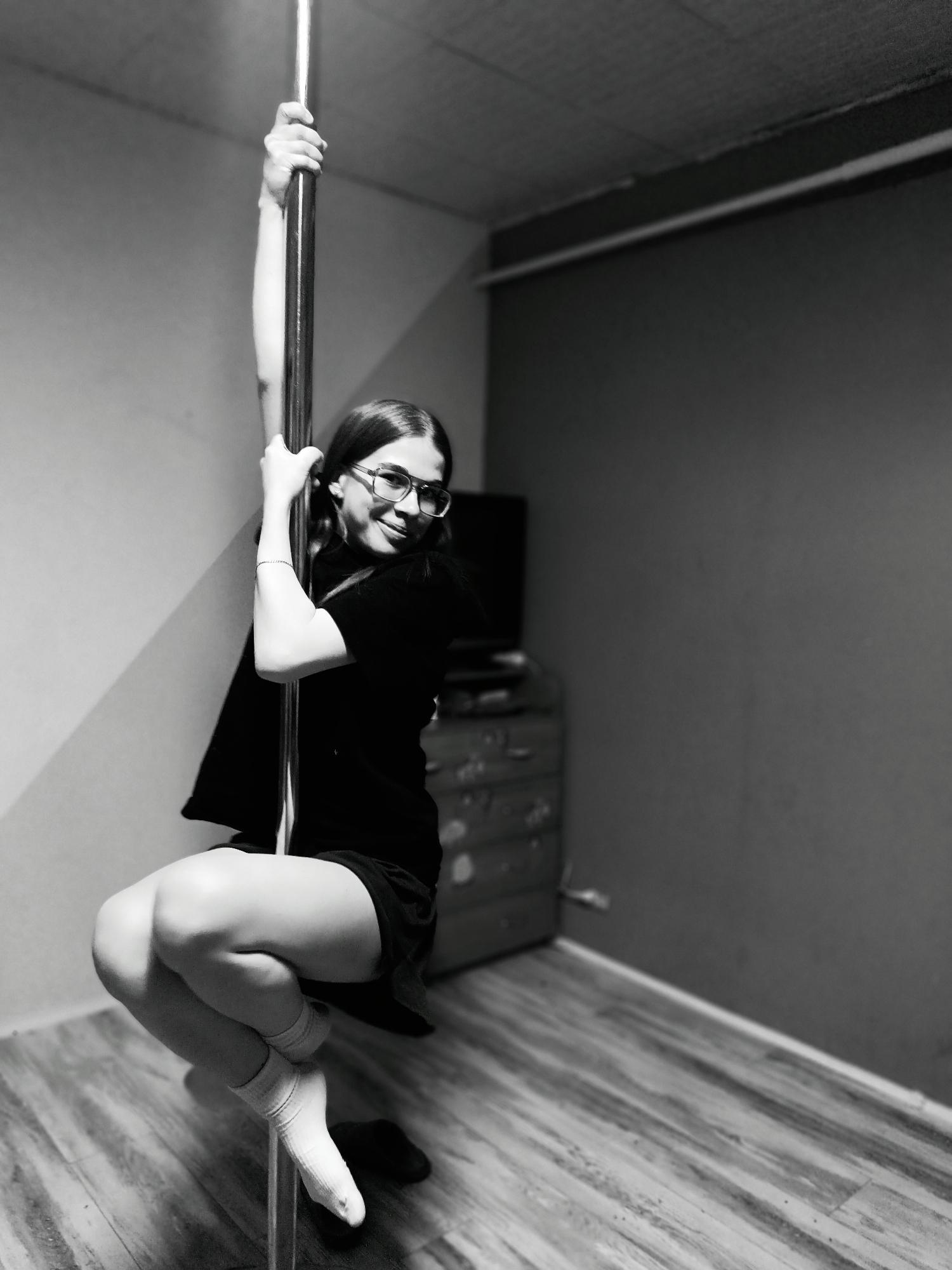 The image shows a young woman posing on a metal pole in what appears to be an indoor setting. The woman is wearing glasses, a dark t-shirt, and dark shorts, with light-colored socks on her feet. She is gripping the pole with both hands and has her legs bent, with one leg extended to provide support. The room has wooden flooring and plain walls, with a dresser and a television set visible in the background. The lighting is soft, casting shadows that highlight the pole and the woman. The exact location within the city of Unknown, Unknown cannot be determined based on the information provided in the image.