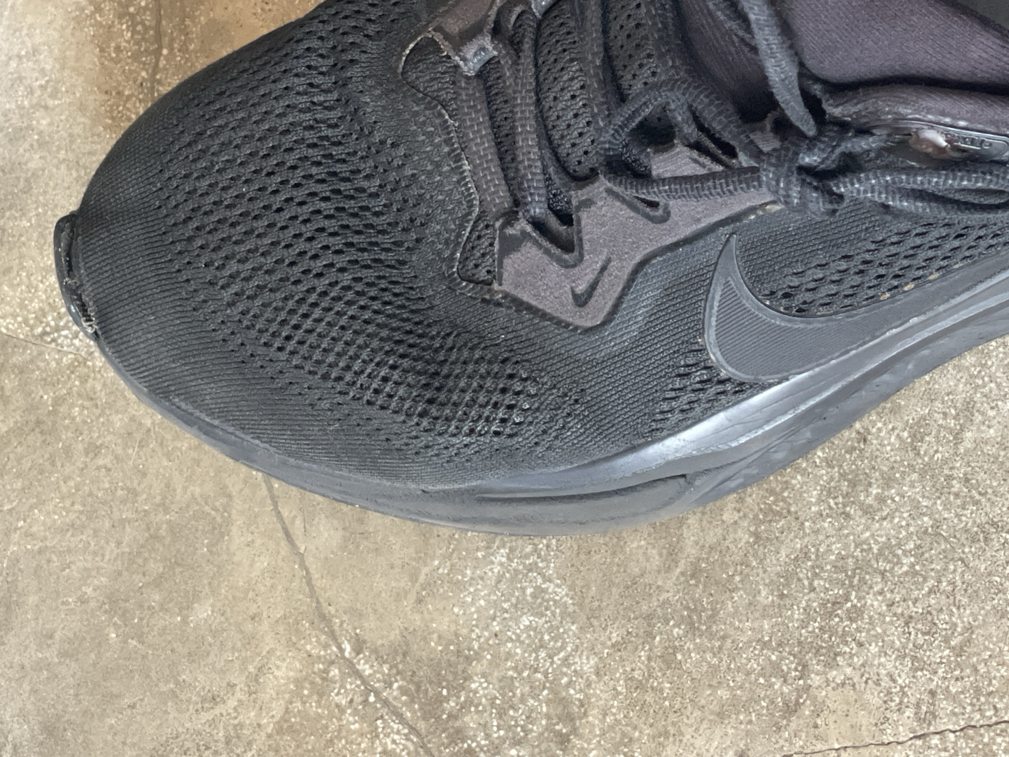 A close-up shot shows a black running shoe on a concrete surface. The shoe has a mesh upper, black laces, and a black Nike swoosh. The concrete surface is light tan with small white specks. The image does not provide any information to determine the location or connection to North Kuta, Indonesia.
