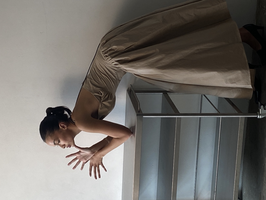 An adult female individual is depicted indoors, leaning with her left elbow on a stainless steel, four-tiered utility cart. The background consists of a plain, light-colored wall and a dark floor. The precise location within the city of London, United Kingdom, is not discernible from the image. The subject is in profile, facing left, with dark hair tied in a ponytail. She wears a light brown or beige strapless dress with a full skirt and black high-heeled shoes. Her hands are raised near her face, fingers splayed outwards. A bracelet is visible on her left wrist, and a thin, dark strap extends over her right shoulder. The utility cart, equipped with wheels and a right-side handle, has empty shelves.