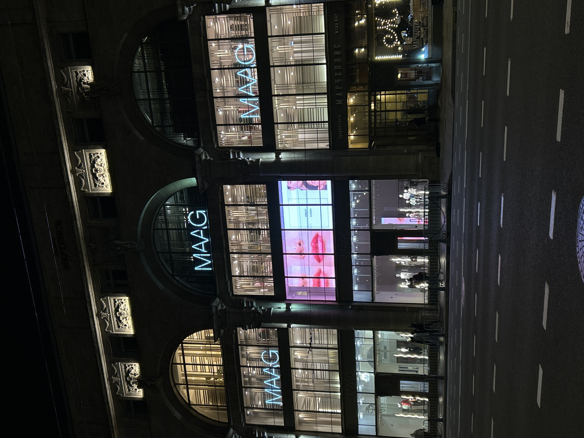 This image shows the MAAG department store in Saint Petersburg, Russia, at night. The building has three tiers of large, arched windows, each illuminated with the store's name in neon lights. A large digital screen displays an advertisement between the top and middle windows. The street in front of the store is dark and marked with white lines for lanes. Mannequins are visible in the store windows. There are architectural details on the left side of the building.