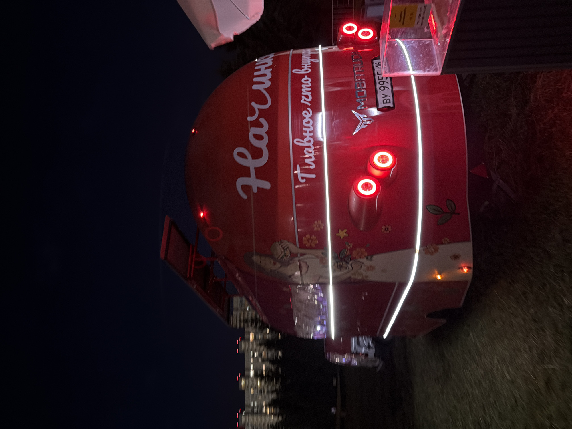 The image captures a red food truck at night in an outdoor setting. The food truck is adorned with white lettering that reads "Начин" on its upper rear section and "Главное что внутри" below. There is a decal of a woman holding a burger on the side of the truck. The rear of the truck features circular red taillights and a license plate that reads "BY 9955 16." A logo for "MOSITRUCK" is also visible on the rear. In the background, there are blurred lights of buildings and trees, suggesting an urban or suburban environment. A portion of a white umbrella and a glass display case are visible on the right side of the frame. The ground is covered in grass.