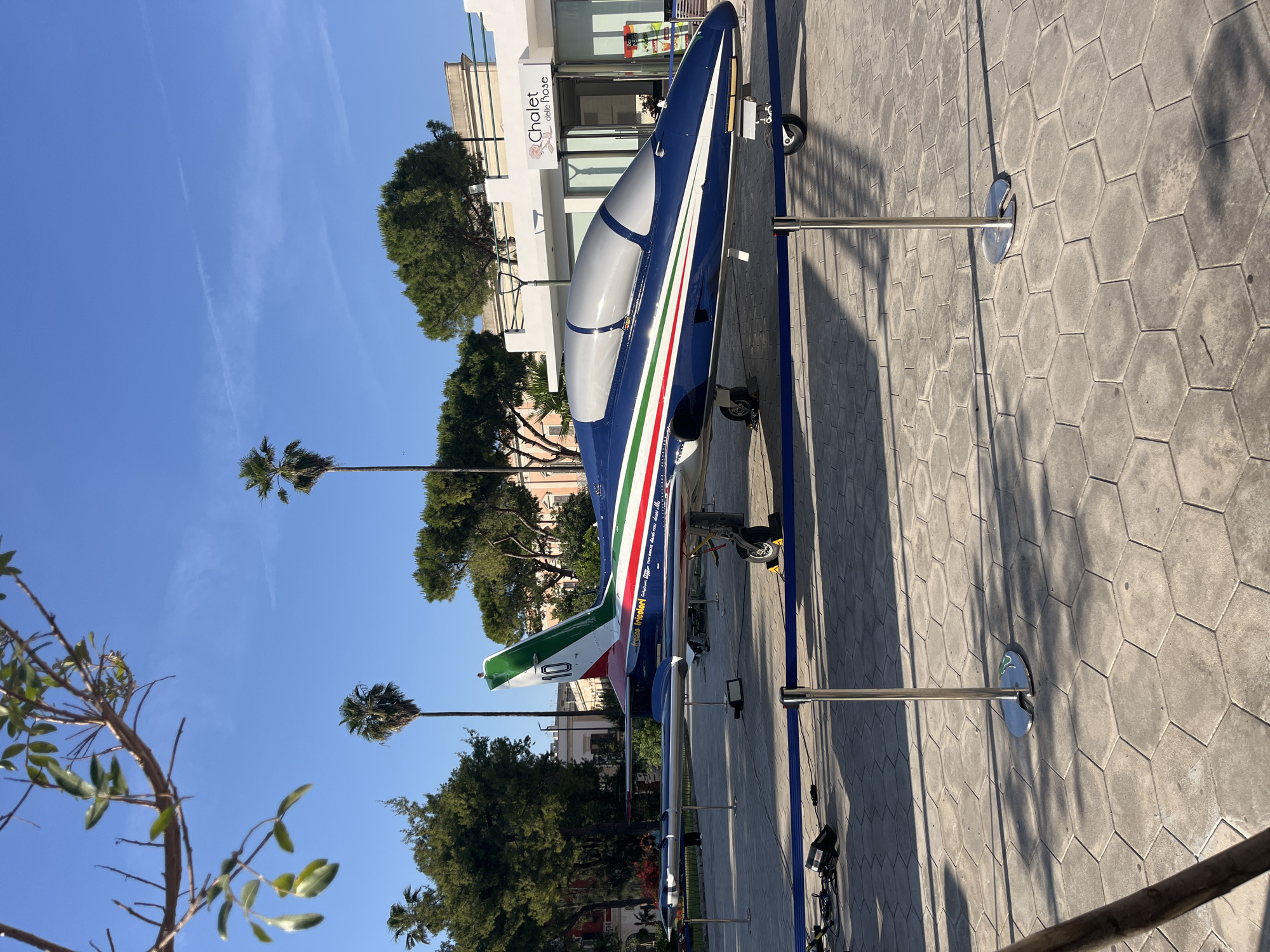 The image captures an Aermacchi MB-339PAN, a jet aircraft used by the Frecce Tricolori, the aerobatic demonstration team of the Italian Air Force, displayed on a hexagonal-patterned paved area in Galatina, Italy. The aircraft is painted in the Frecce Tricolori's distinctive blue, white, and green livery. It is mounted on metal stands, and a blue rope barrier surrounds it.
Behind the aircraft, there are trees, a building with a sign that reads "Chalet delle Rose", and clear blue sky with palm trees.
The scene is bathed in sunlight, casting shadows across the pavement.