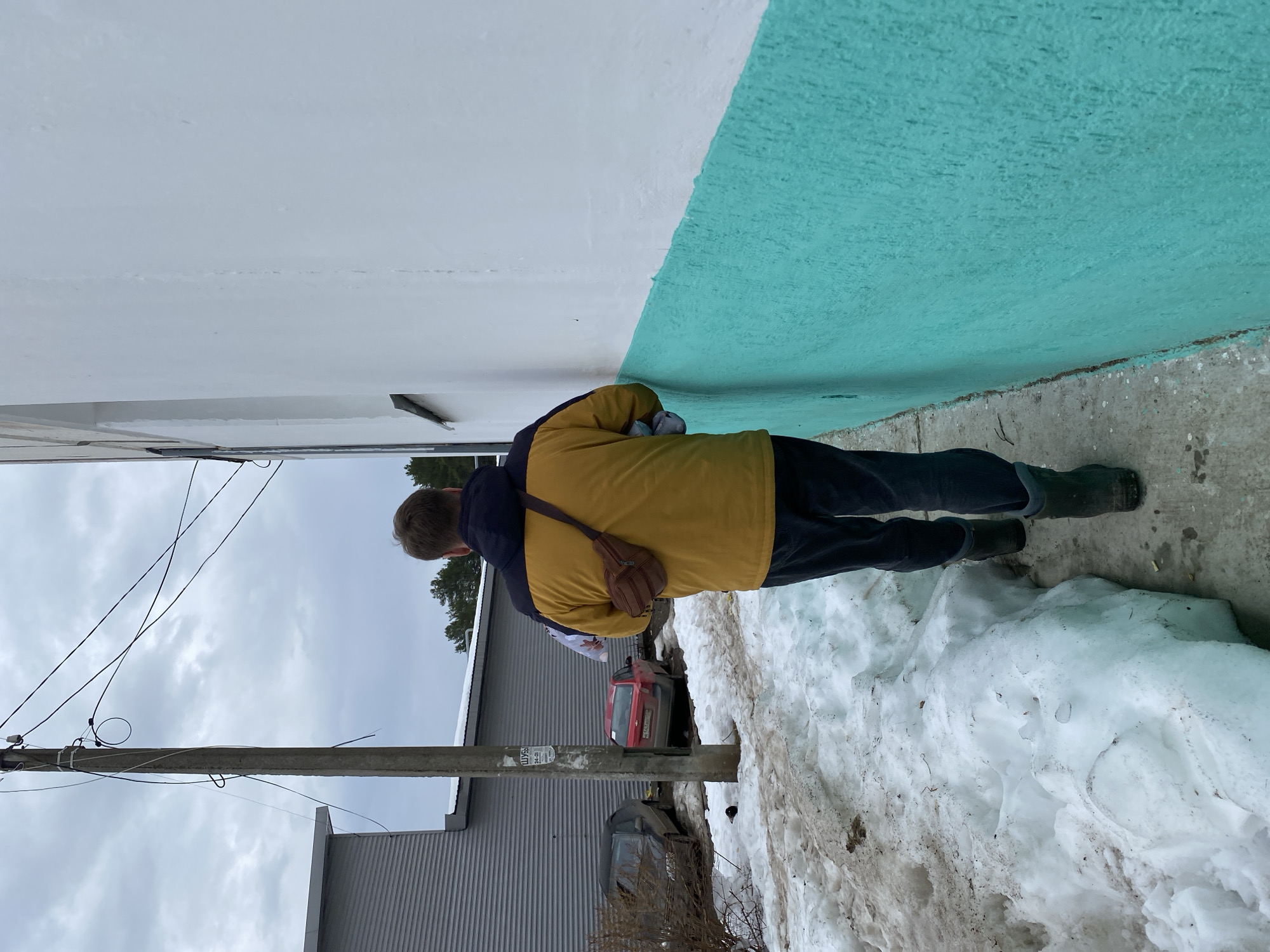 A man in a yellow jacket and dark pants stands beside a building with a white top half and a blue-green lower half. He has a brown messenger bag slung over his shoulder. The ground beside the building is covered in snow. A pole with wires runs vertically in the left side of the frame. In the background are other buildings and bare trees, partially obscured by the pole and wires. A red vehicle is visible between the man and a building in the background. The location is Chaikovsky, Russia.