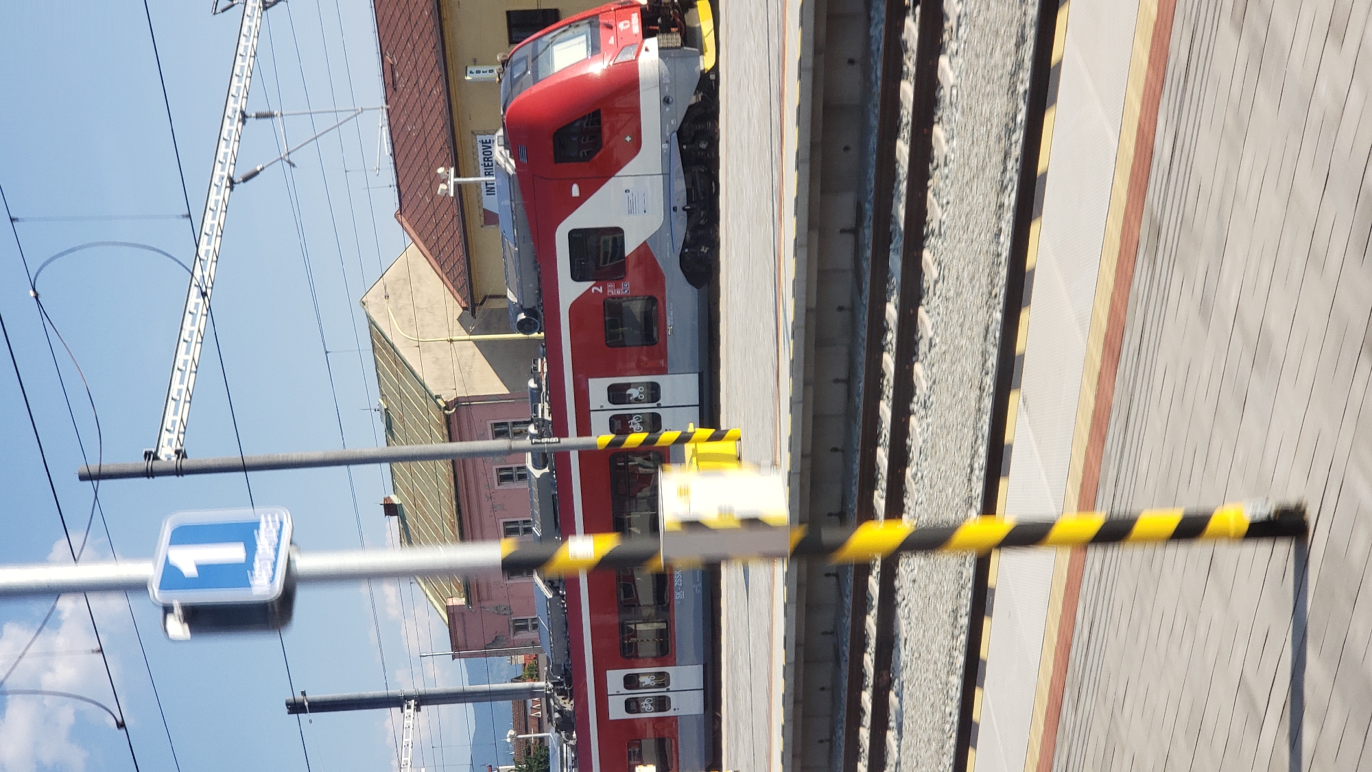 The image depicts a red and white passenger train stopped at a platform in Žilina, Slovakia. The train is positioned on tracks, with the platform in the foreground. A sign with the number "1" is visible on a pole in the foreground, indicating Platform 1. In the background, buildings with reddish-brown and light-colored facades can be seen, along with overhead power lines for the train. The sky is clear and blue. The scene appears to be a typical railway station environment.
