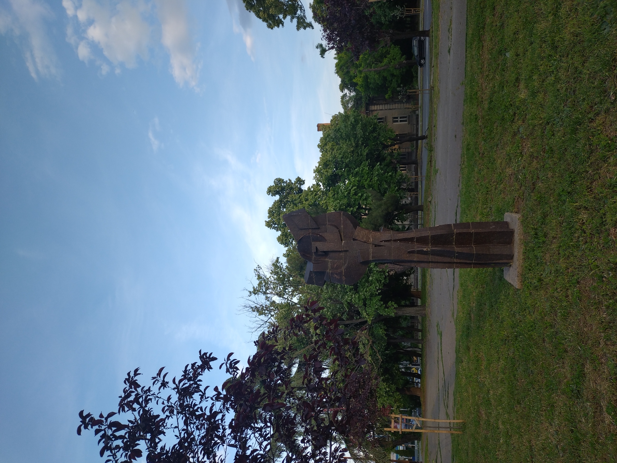 The image shows a statue in Stara Zagora, Bulgaria. The statue is made of dark metal and is located in a park area with trees, grass, and a paved path. A building is visible in the background behind the trees along with some parked cars. The sky is blue with some clouds. A tree with dark red leaves is located in the lower left corner of the image.