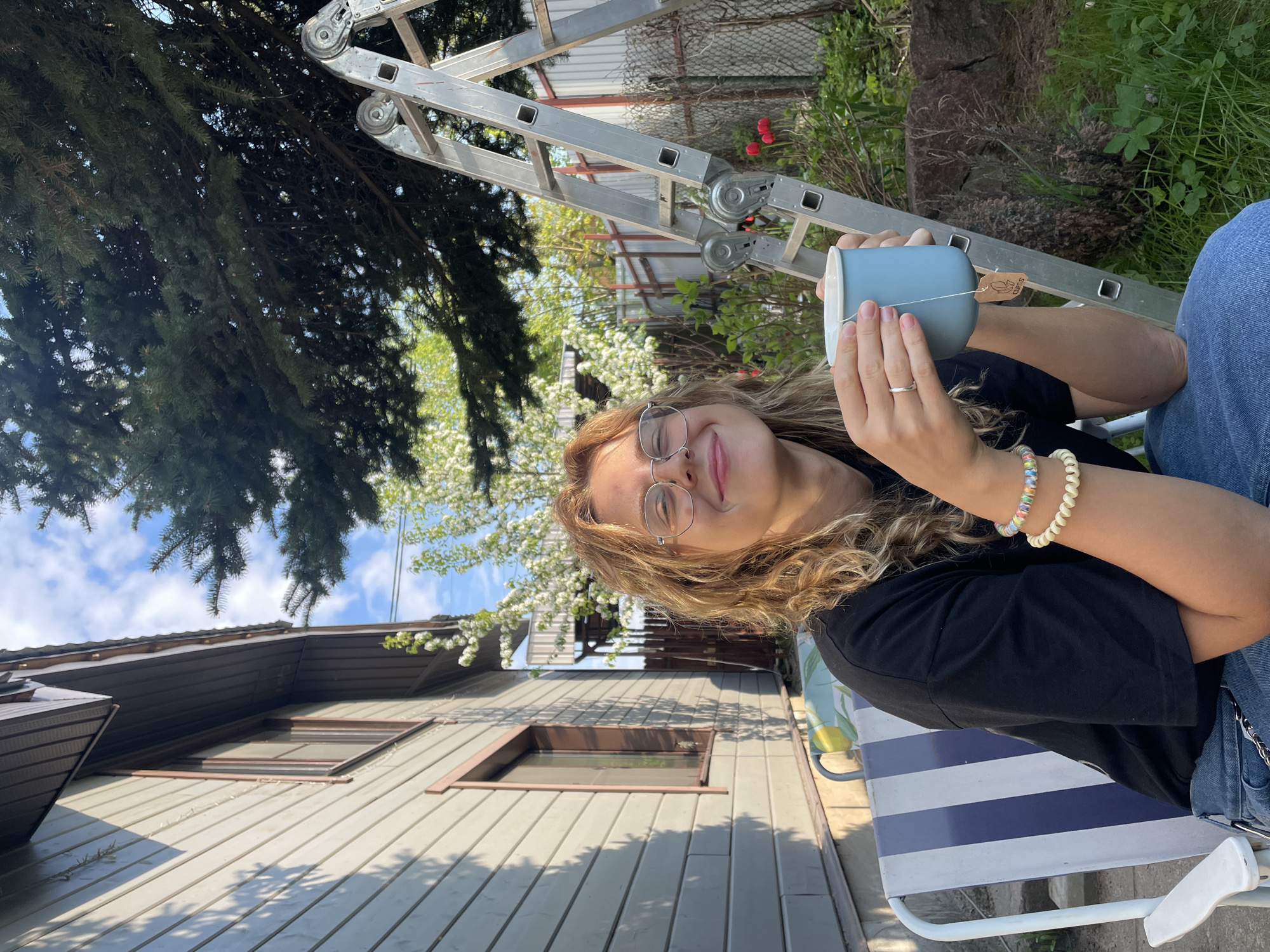 A woman with light brown curly hair and glasses is holding a light blue mug with a tag attached. She is wearing a black shirt and jeans and has bracelets on her left wrist. She is sitting on a blue and white striped chair outdoors. In the background, there is a wooden house with a brown roof, a tree with white blossoms, and a metal ladder. The location appears to be an outdoor area in Sestroretsk, Russia, based on the architectural style of the house and typical flora.