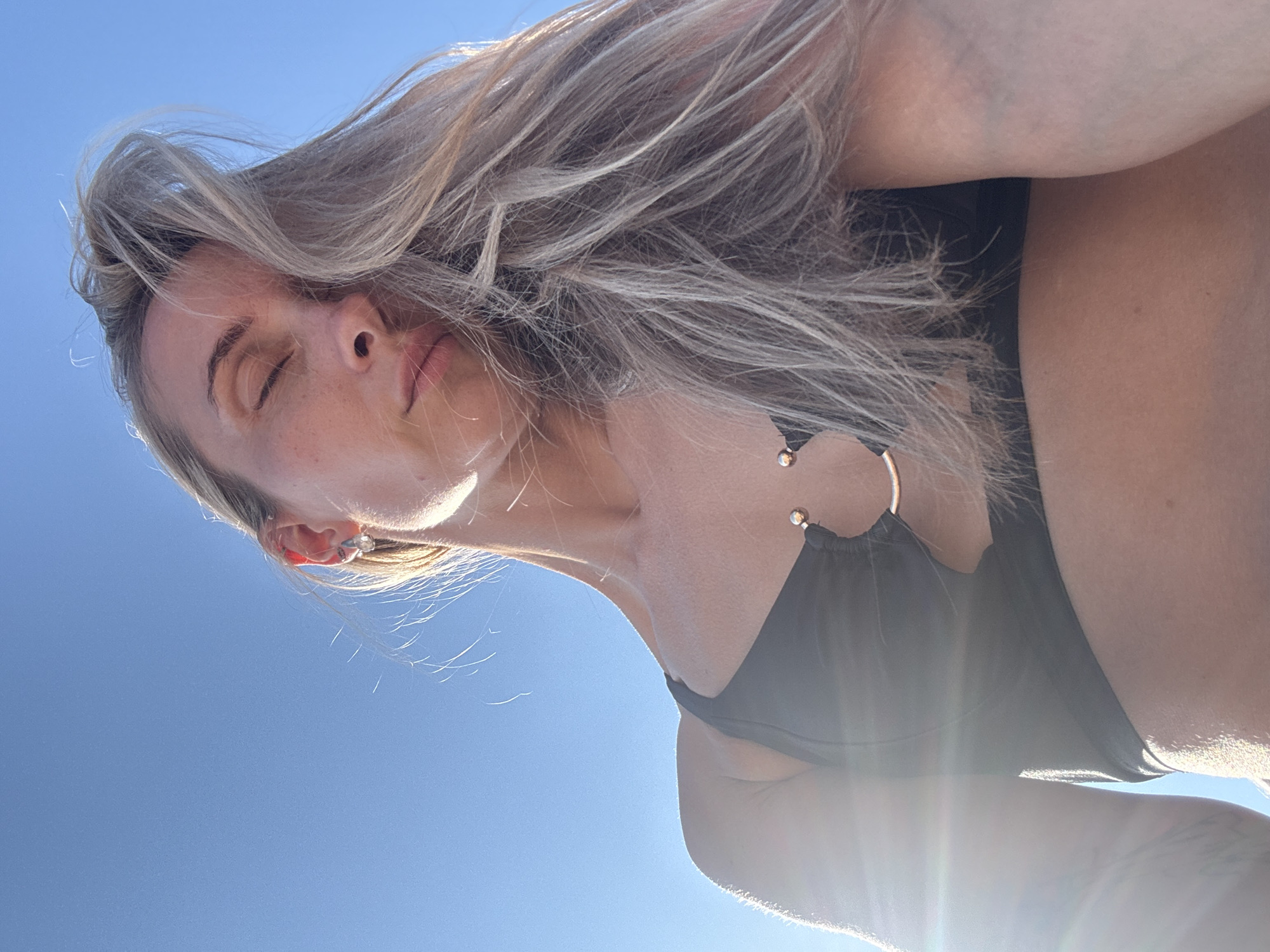A woman with blonde hair is captured in a low-angle shot, looking upwards with her eyes closed. She is wearing a black swimsuit with a circular metal accent. The bright sunlight casts a lens flare across the image, suggesting it's daytime. The background is a clear, vibrant blue sky, indicating fair weather. The woman's face is tilted, with her hair flowing. The scene is outdoors, likely in a location with a clear sky, possibly a beach or an open area. The overall mood appears relaxed and serene, accentuated by her closed eyes and the peaceful sky. There is no visible text in the image.