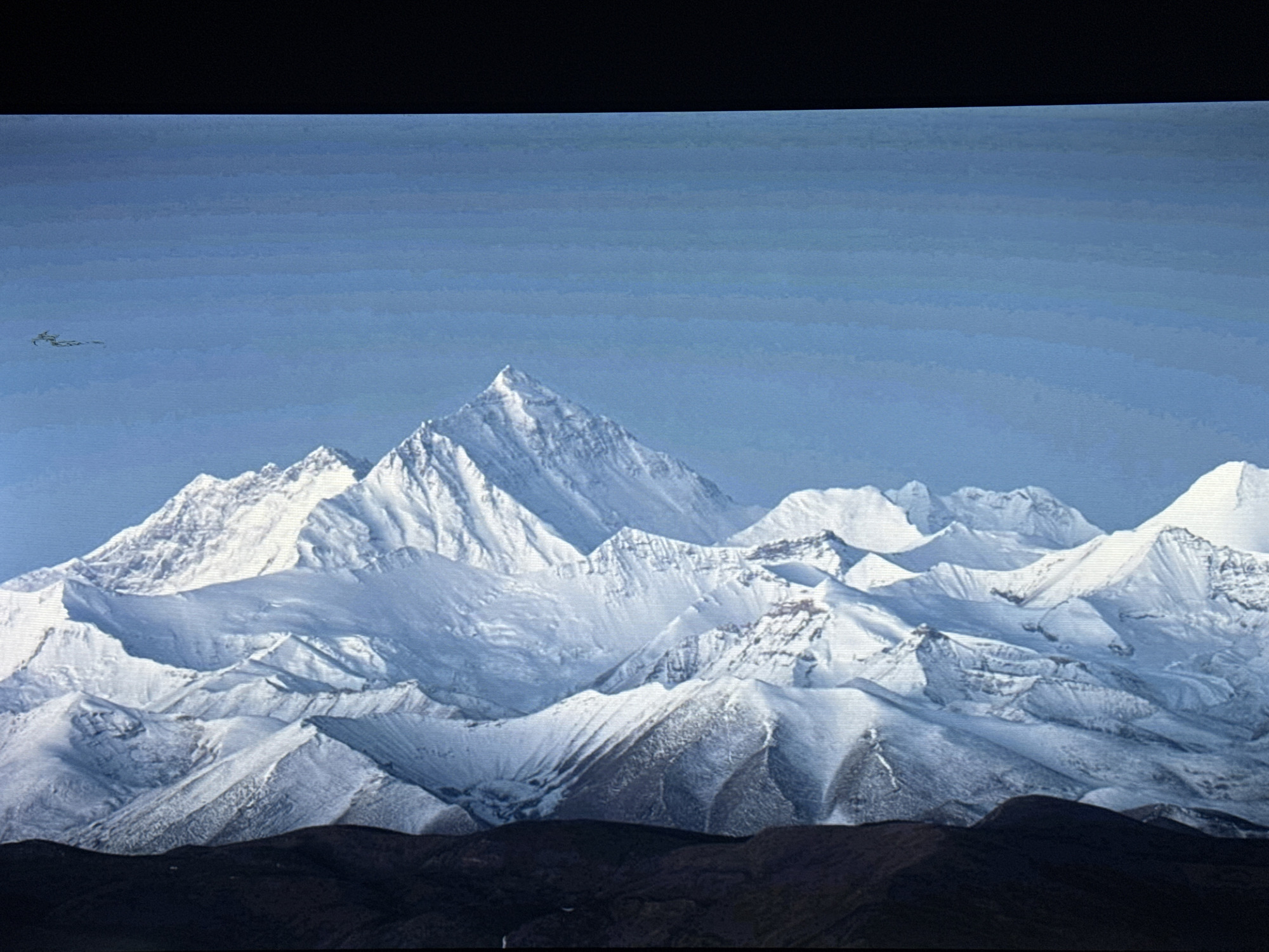 The image depicts a majestic mountain range covered in snow under a clear, pale blue sky. The peaks are sharp and rugged, with deep shadows indicating dramatic relief. The lower foreground consists of dark, barren hills, contrasting sharply with the snow-capped mountains above. There is no visible presence of the city of Жәпек батыр ауылы, Kazakhstan, in this image. A small, dark object, possibly a bird, is visible in the upper left portion of the sky, appearing as a distant speck. The overall impression is one of vast, natural grandeur and isolation.