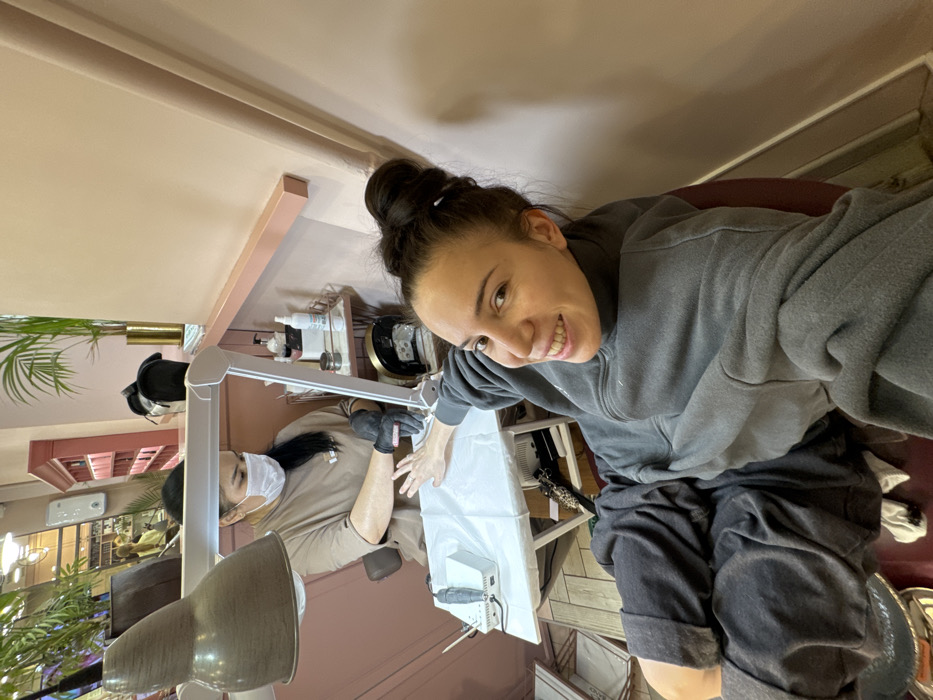 This image depicts an indoor scene within a nail salon located in Moscow, Russia. The primary subject, a female client, occupies the foreground, facing and smiling at the camera. She has dark hair styled in a high bun and is wearing a dark grey hooded sweatshirt and dark grey sweatpants, seated in a reddish-brown chair. Her left hand is extended towards a nail technician.
The nail technician, positioned in the background, is a female wearing a light brown top, a white face mask, and black gloves. She is seated opposite the client, actively performing a manicure service on the client's left hand. Her workspace includes a small white table covered with a disposable white mat. On the table, a white rectangular nail drill machine is visible. An adjustable magnifying lamp with an integrated ring light illuminates the work area, accompanied by a larger overhead lamp with a brown metallic shade. A black arched UV/LED nail lamp is positioned near the client's hand.
Adjacent to the technician's station is a small, gold-framed tiered trolley holding various bottles and containers, likely nail products. The salon environment features light peachy-pink walls, a prominent pink decorative railing running along a wall, and several green potted plants. Reflections in the upper left background suggest additional salon spaces with shelving and lighting fixtures. A white wall-mounted dispenser is also visible. The floor appears to be a light-colored wood.