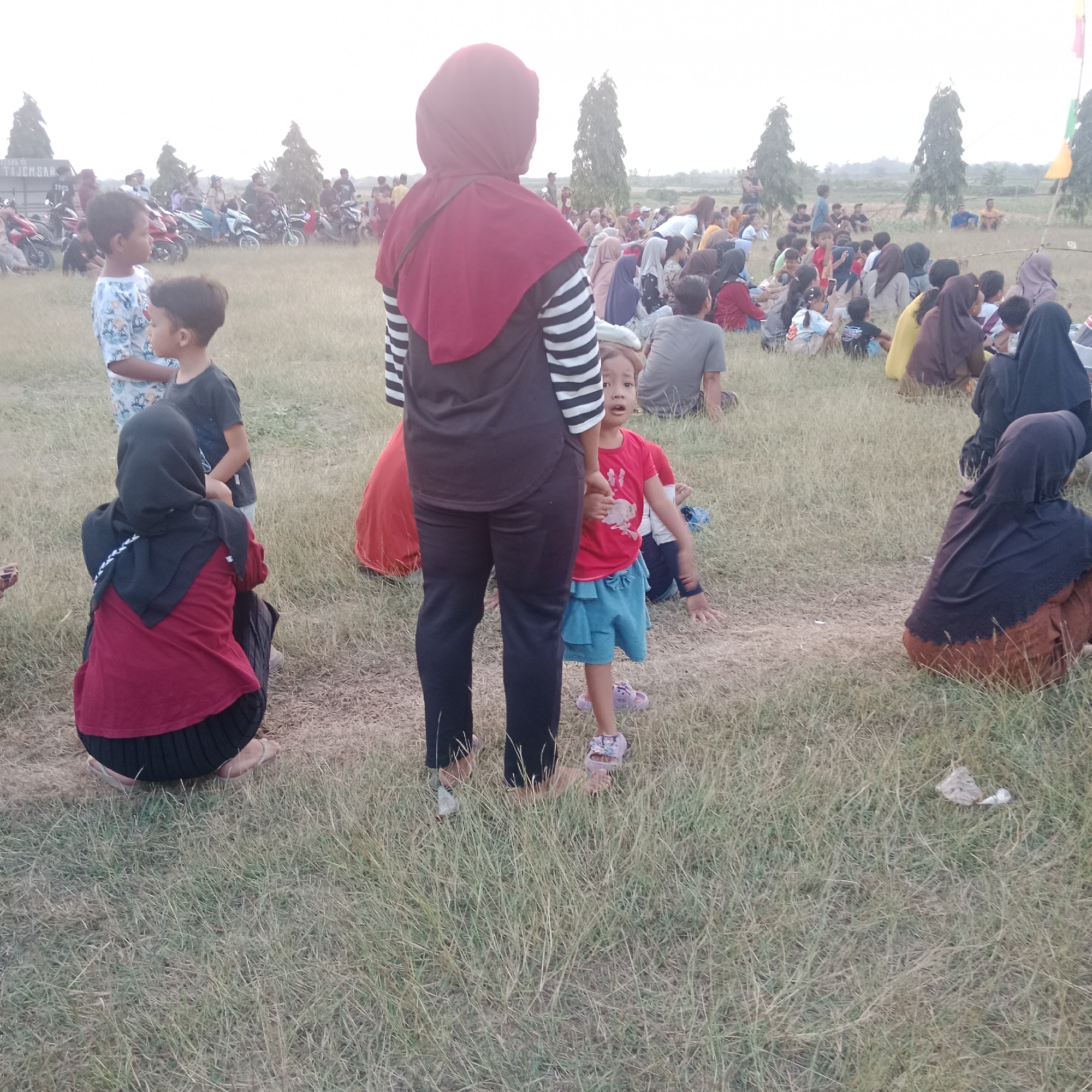 The image depicts an outdoor gathering of people, predominantly in a grassy field, with some vehicles, likely motorcycles, visible in the background. The scene suggests a community event, possibly a festival or informal meeting, taking place in Tegowanu Wetan, Indonesia. Several individuals are seated on the ground, some are standing, and a few children are present. The attire of the people includes traditional Islamic head coverings, such as hijabs, worn by many women. The environment appears somewhat arid, with dry grass and a light-colored sky. The overall impression is one of a casual gathering in a rural or semi-rural setting.