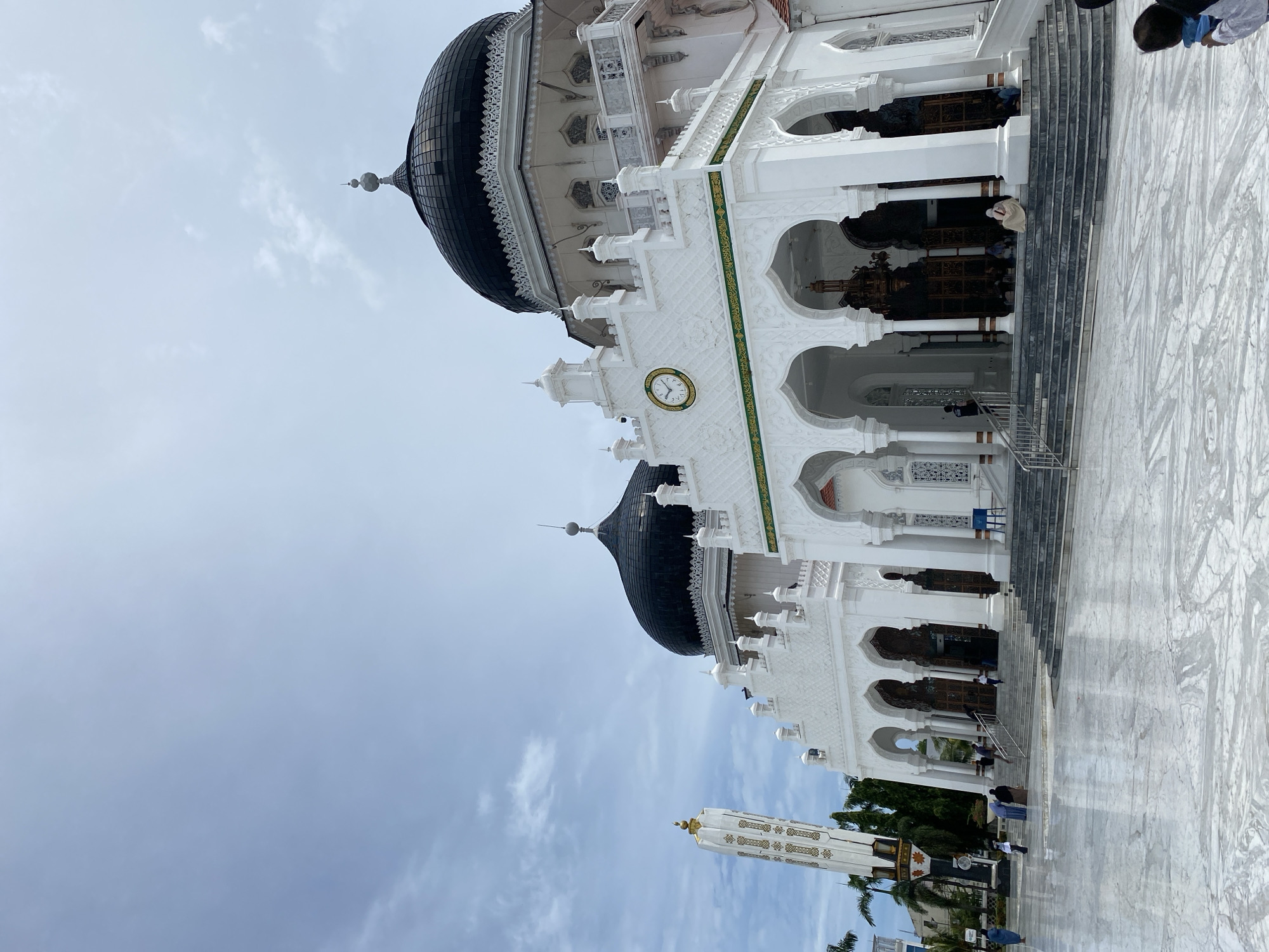 The image shows the Baiturrahman Grand Mosque in Banda Aceh, Indonesia. The mosque is white with black domes and intricate architectural details. A clock is visible on the facade. Several people are present, some are walking or standing, while others appear to be praying. The environment includes a paved exterior and a partly cloudy sky.