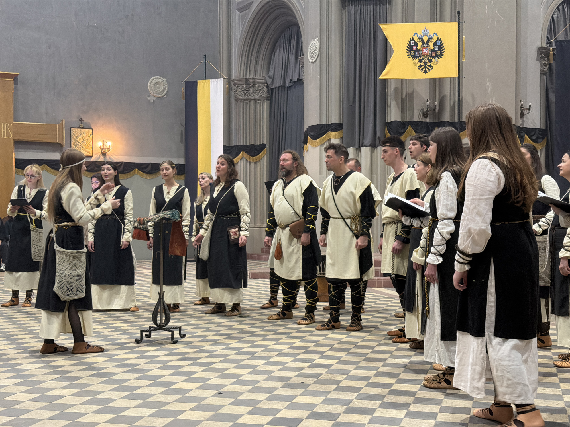 A large group of individuals, appearing to be a choir or vocal ensemble, stands in a spacious, high-ceilinged indoor setting, likely a historical hall, church, or cathedral. The floor is a distinct black and white checkered pattern.
The subjects are dressed in period-inspired costumes, predominantly featuring black and off-white or cream colors. Both men and women wear long tunics or dresses, often with dark outer garments over lighter undergarments. Many of the men have their legs wrapped with yellow or dark cords, and all wear simple, flat sandals or shoes. Several individuals hold open books or music sheets, and some have open mouths, indicating they are singing or about to sing.
Two historical Russian Imperial flags are prominently displayed. On the right, a large yellow flag features a black double-headed eagle emblem. On the left, another flag displays vertical stripes of black, yellow, and white. The architectural elements include tall arches, columns, and decorative moldings, suggesting a grand, historical building. A wall-mounted light fixture and an icon or relief are visible on the left wall. A decorative metal stand is positioned in the foreground.
The activity taking place is a musical performance or rehearsal, likely of historical or traditional music, given the attire and setting. The performers appear focused and engaged. On a wooden structure to the far left, partially visible letters "HS" can be seen, possibly part of a larger inscription. The indoor lighting is artificial and provides a somewhat subdued atmosphere. The presence of the Russian Imperial flags strongly hints at a Russian context, aligning with the provided location of Saint Petersburg, Russia.