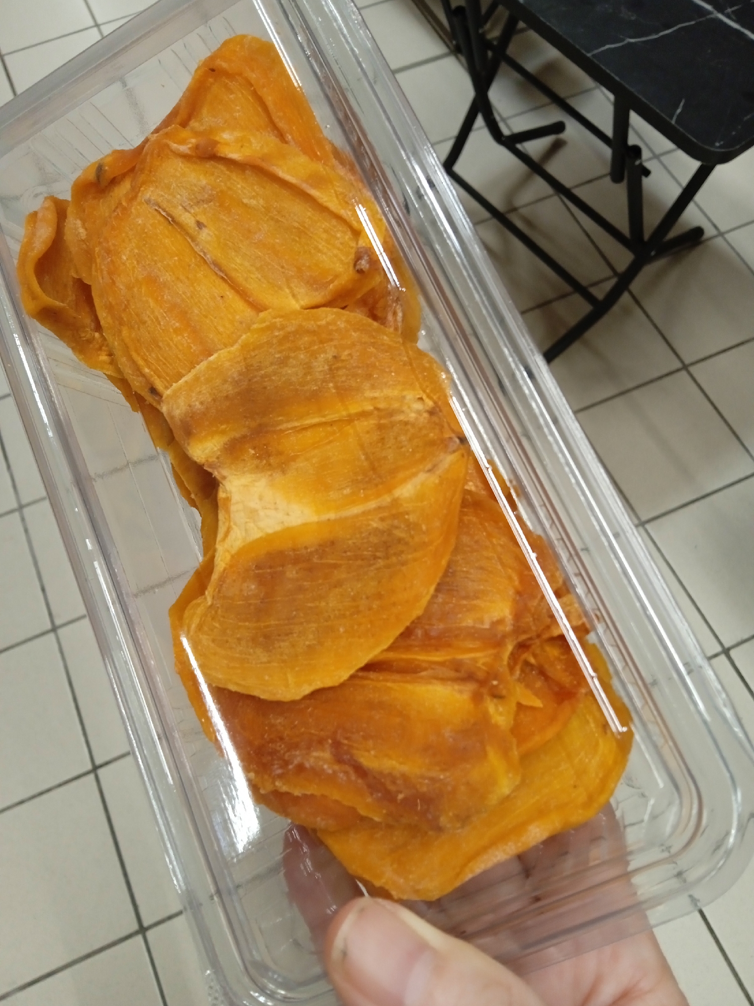 A clear plastic container of dried persimmon slices is held in a person's hand. The dried persimmons are orange and appear somewhat leathery. The container is resting on a tiled floor. In the background, a black table with a marble-like pattern is visible. The overall scene is an indoor environment, likely a store or market, in the city of Yenişehir, Turkey.
