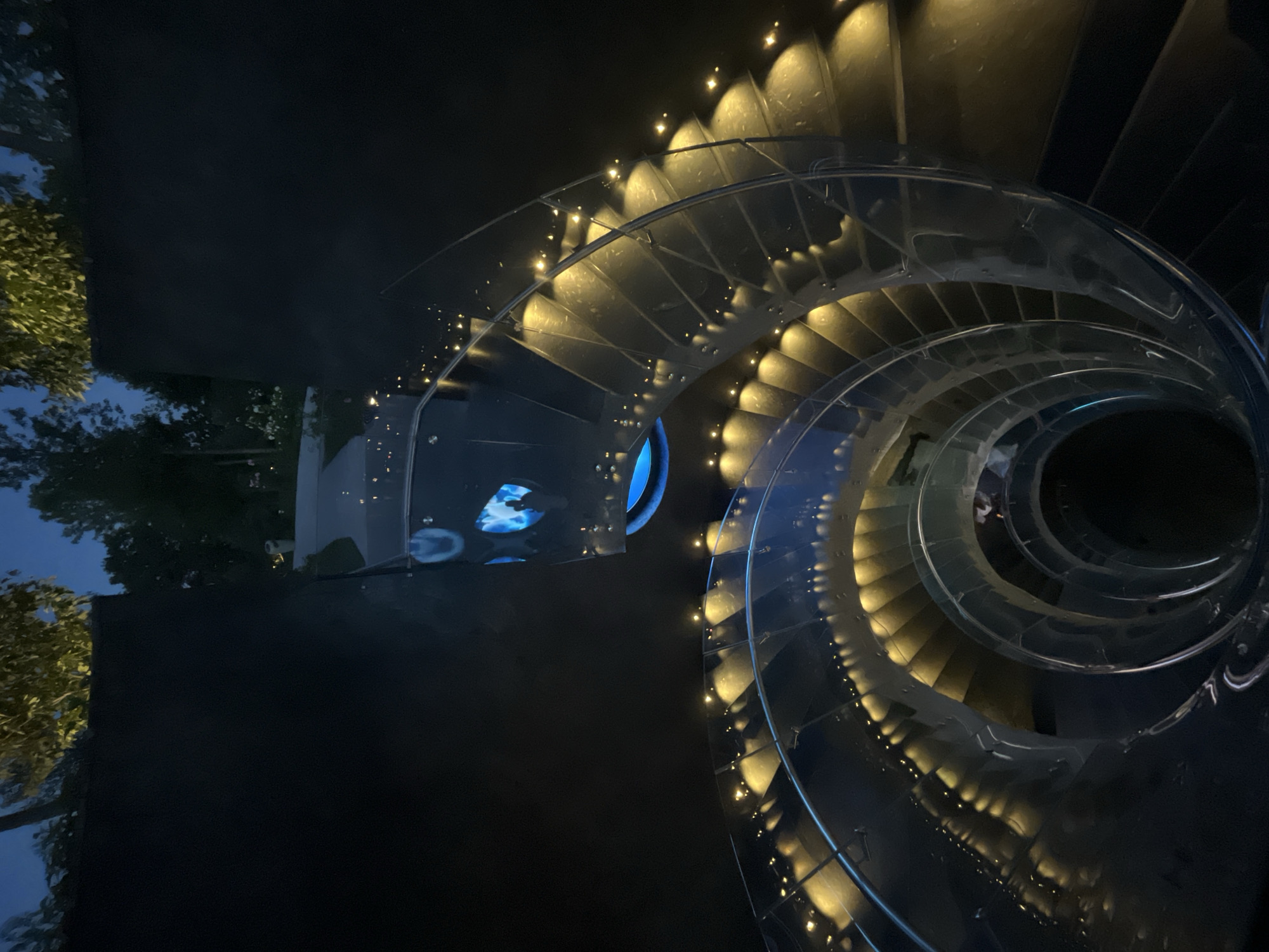 This image depicts a spiral staircase at night, illuminated by warm yellow lights embedded along its railing and steps. The staircase appears to be made of glass and metal, creating a modern and elegant aesthetic. The ambient light suggests it is either dusk or night, with a dark, possibly outdoor environment surrounding the structure. In the background, faint outlines of trees are visible against a dark blue sky. The lighting creates a dramatic and almost ethereal atmosphere. There is no visual information in the image that indicates a specific location within the city of Krasnodar, Russia.