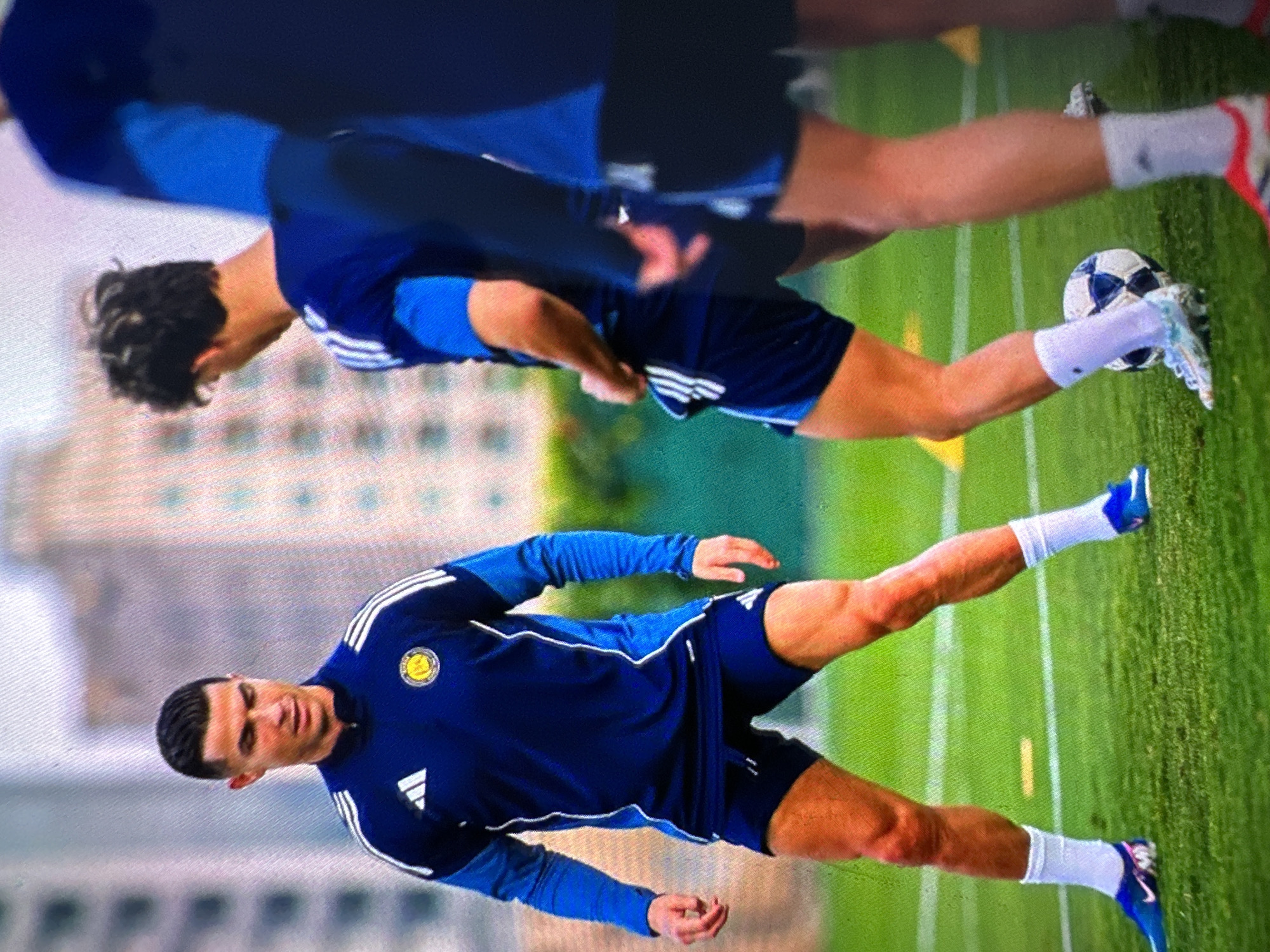 This image shows Cristiano Ronaldo on a soccer field, wearing a dark blue Adidas tracksuit and shorts. He is in the foreground, with two other blurred figures in the background, also in soccer attire. The field is green with white lines, and in the background, there are blurred buildings that suggest an urban environment. The presence of Adidas branding and a soccer ball indicates a sports training or game context. The image's quality is somewhat low, with visible pixelation. The prompt asks for the location in the city of Khimki, Russia, but there are no visual cues in the image to confirm this specific location.