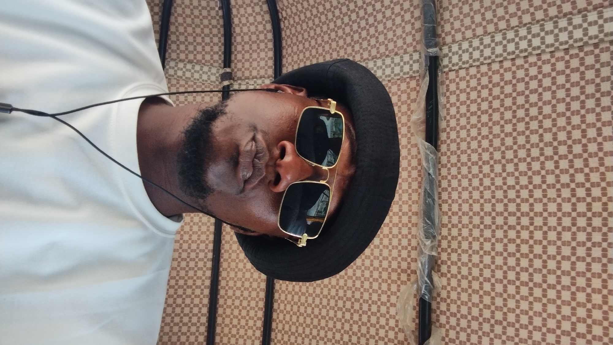 A close-up, slightly rotated image shows a man with dark skin, wearing a black hat and gold-rimmed sunglasses. He is wearing a white shirt and has wired earphones visible. The background is a brown and tan patterned material, likely a mat or fabric. A black metal pole partially wrapped in plastic runs vertically along the right side of the image. The location is not discernable from this image.