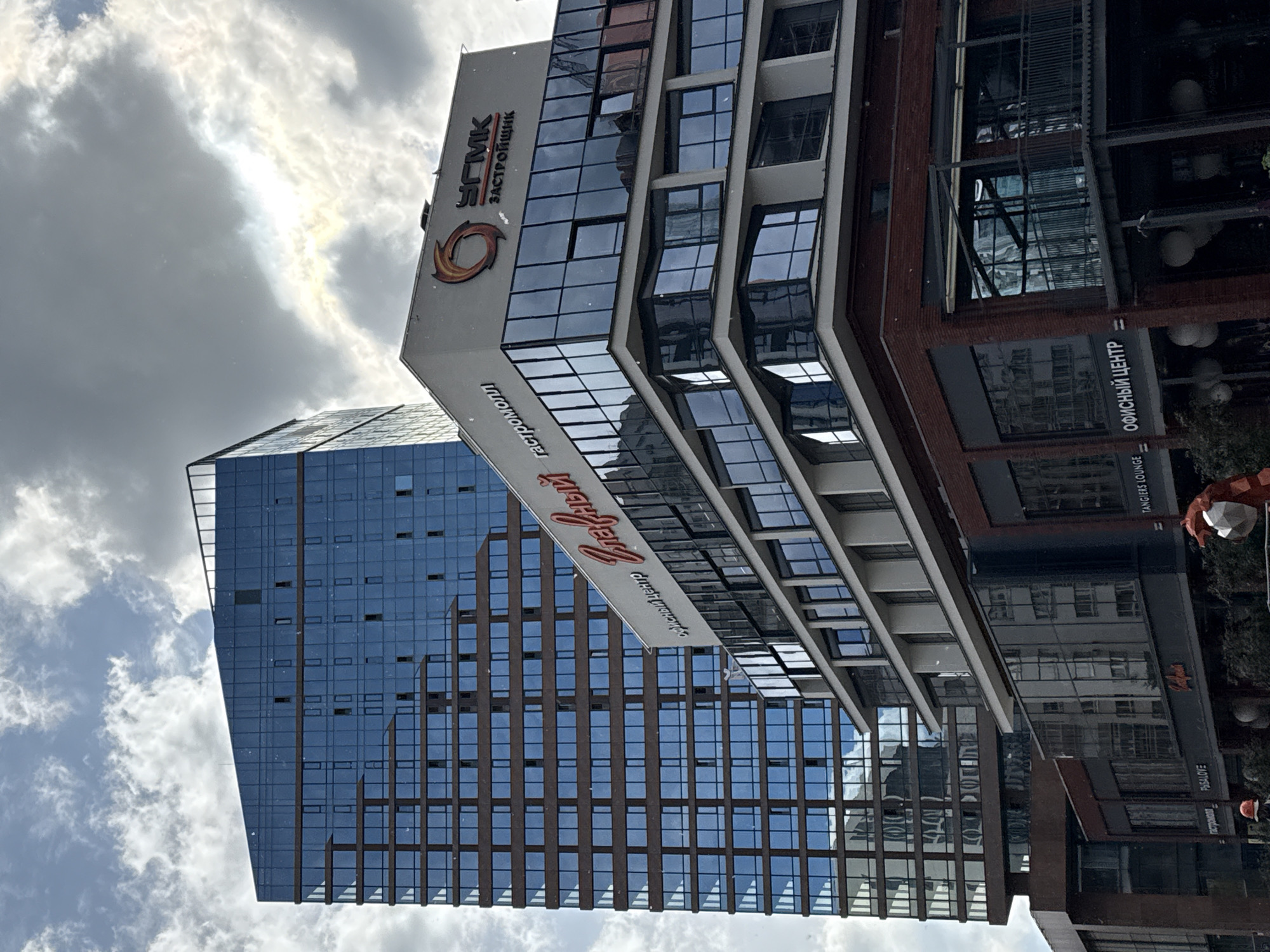 The image shows a view of the "Glava" business center and residential complex in Yekaterinburg, Russia, on a cloudy day. A tall, modern building with a blue glass facade and brown accents is visible on the left. To its right, a white building with a distinctive red spiral logo and Cyrillic lettering is prominent. This building features multiple stories with large, reflective windows and a dark red brick base. In the foreground, lower buildings with commercial signage, including "ОФИСНЫЙ ЦЕНТР" (Office Center) and "TANGIERS LOUNGE", are visible. The sky is filled with dramatic clouds, with patches of sunlight breaking through.