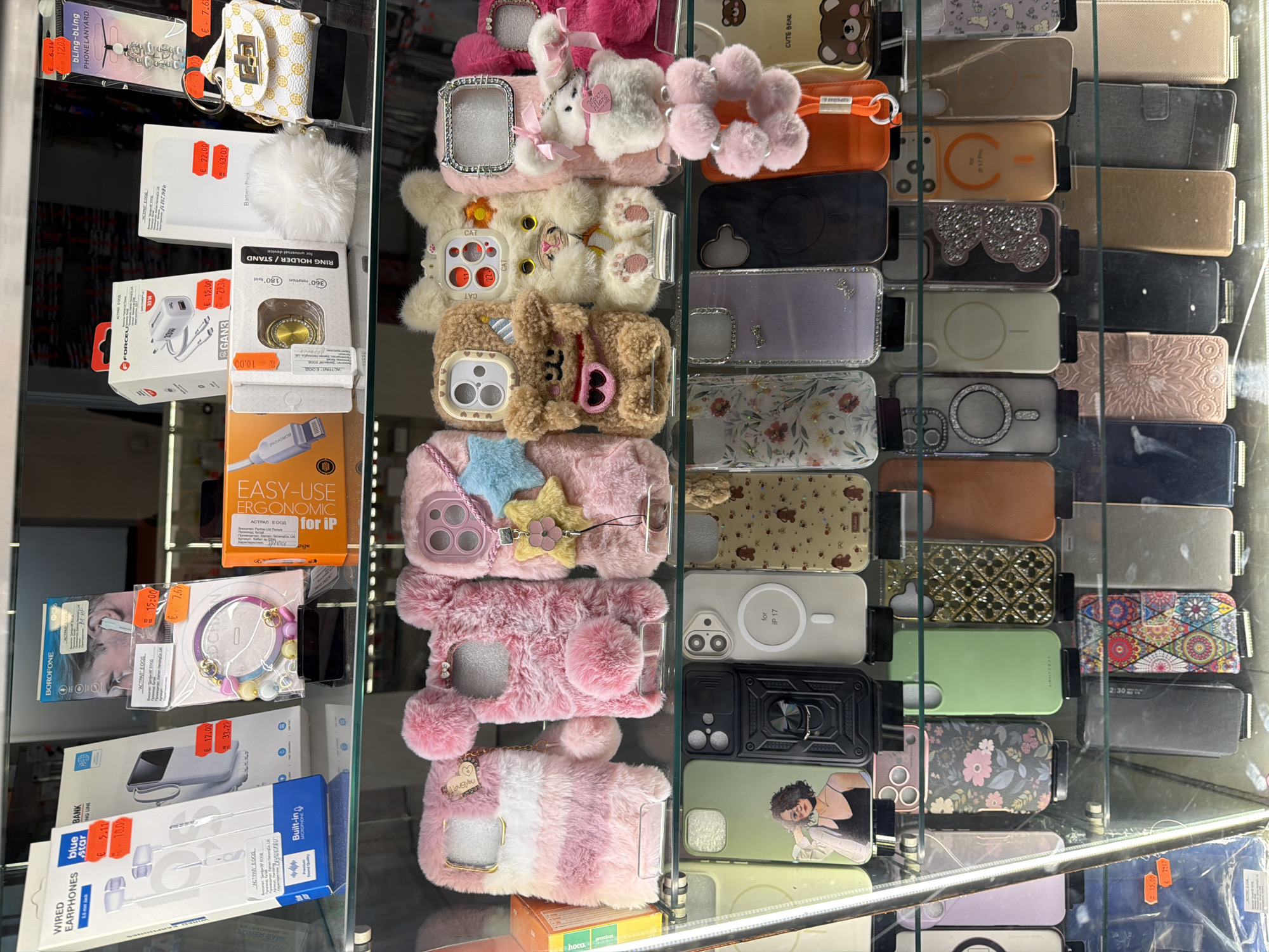 The image depicts a retail display shelf in a shop located in Burgas, Bulgaria. The display showcases various phone accessories, primarily phone cases and some electronic items. The items are arranged on multiple shelves, with a clear emphasis on colorful and decorative phone cases.
On the top shelf, several boxed electronic accessories are visible, including headphones, chargers, and portable batteries. Among these, there are also decorative items like a bracelet and a small handbag-shaped item. Pricing tags are visible on some of these products.
The middle shelf is dominated by an assortment of fluffy and decorative phone cases, many featuring animal motifs like bears and cats, and vibrant colors like pink and white. There are also several cases with embroidered patterns and embellishments.
The bottom shelf displays a variety of phone cases, some with patterned designs, clear cases with embedded glitter, and solid colored cases. A few other electronic accessories and phone chargers are also present on this shelf.
The overall environment appears to be a well-lit retail store. The display is orderly, with each item presented for sale. The presence of pricing information suggests a commercial transaction environment.