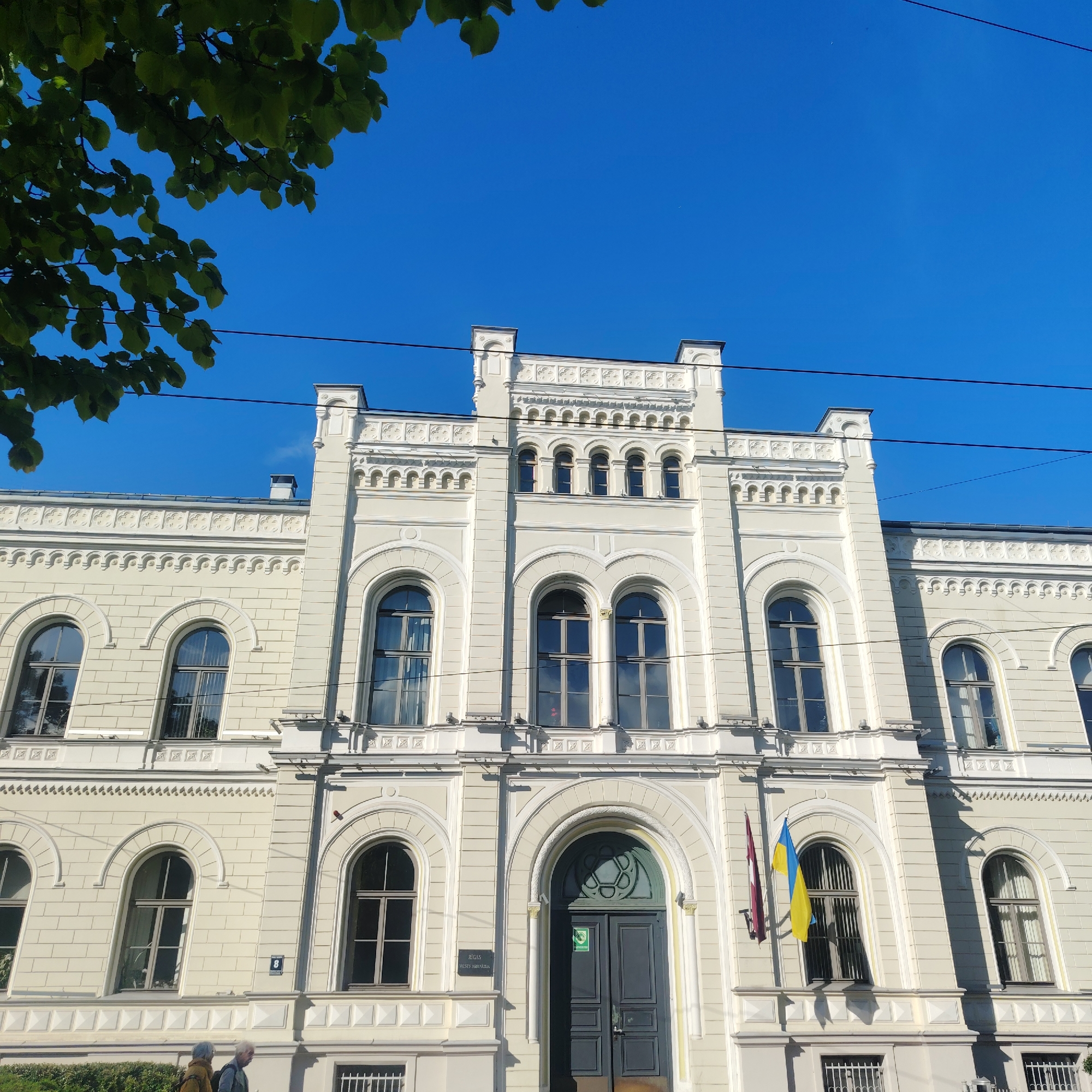 A building stands prominently against a clear blue sky. The architecture is light-colored, featuring arched windows and ornate detailing. A tree branch with green leaves is visible in the upper left corner, partially obscuring the sky. Two flags, one Latvian and one Ukrainian, are mounted to the building's facade on the right of the entrance. The building’s entrance is a large, gray, double door, above which is an arched window filled with a circular design. At the building's base, two people are partially visible, walking from left to right. "Riga Ministry of Foreign Affairs". "Number 8".
