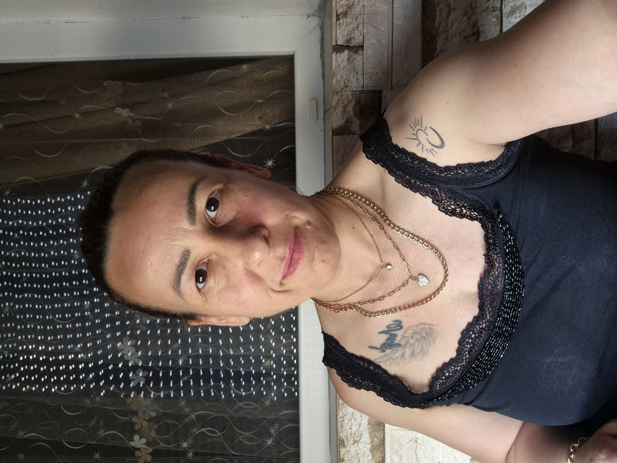 This media file is a portrait of a woman. She is looking towards the camera with a slight smile. She has dark hair pulled back, dark eyes, and light pink lipstick. She is wearing a black, sleeveless top with a lace trim. She has multiple gold necklaces on, one with a heart-shaped pendant and another with a small diamond. She has two tattoos visible: one on her upper right arm with a sun-like design, and a larger tattoo on her chest, appearing to be wings and possibly text. The background shows a window with sheer curtains to her left and a textured wall to her right. The lighting suggests it is daytime.