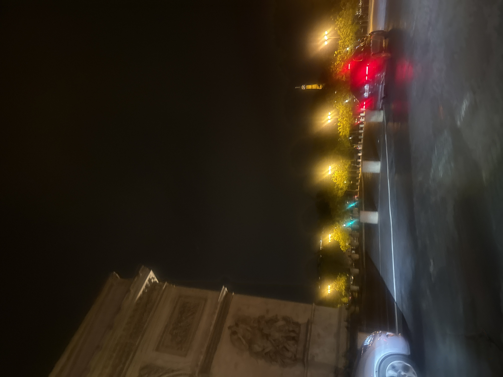The image captures a night scene in Paris, France, featuring the Arc de Triomphe in the lower left corner and the Eiffel Tower visible in the background, further down the avenue. The scene is lit by streetlights illuminating the Champs-Élysées and surrounding area. The street is wet, reflecting the lights. Several cars are also on the street. The scene is at night.
