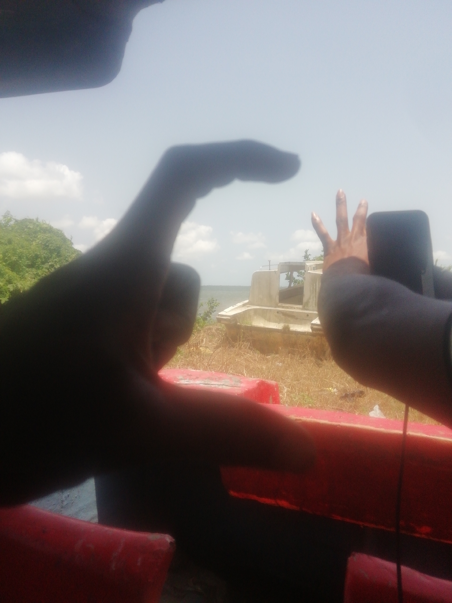 A photograph taken from a boat in Ikorodu, Nigeria, depicts a scene where an individual is using a mobile phone to capture an image of a derelict boat ashore. The image is obscured by the foreground, with the left hand partially obstructing the view and the right hand holding a phone, presumably to take the picture. In the background, an abandoned boat structure rests among dry vegetation near a body of water under a bright sky. The foreground reveals the interior of the boat from which the photograph is being taken, showing red paint and dark interior elements.