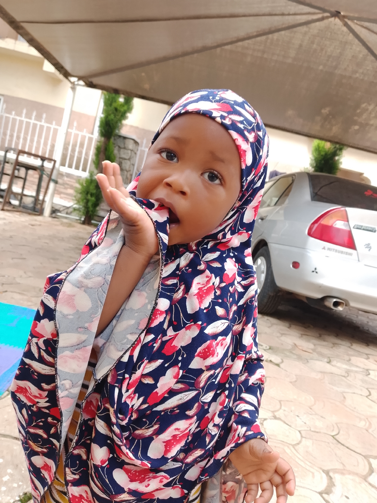 In the city of Jos, Nigeria, a close-up, outdoor, medium shot captures a young child wearing a blue, white, and red floral hijab. The child's left hand is raised to their mouth, and their gaze is directed slightly upwards and to the left. Behind the child, a silver Mitsubishi Lancer sedan is parked on a paved surface. The background includes a covered patio area, some white fencing, and sparse greenery. The lighting suggests it is daytime.
