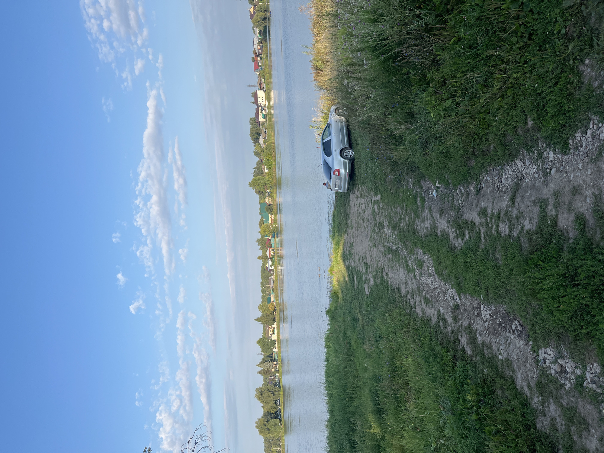 A wide shot captures a serene landscape featuring a calm body of water, likely a river or lake, bordered by a shoreline with lush green grass and a dirt path leading down to the water's edge. On the right side of the frame, tall grasses and wildflowers sway gently. In the middle ground, a silver car is parked on the grassy bank near the water. Across the water, a village or town is visible, with houses of various colors and sizes nestled among trees. The sky above is a bright blue, adorned with scattered white clouds, suggesting a pleasant day. The overall scene evokes a sense of rural tranquility, typical of a location such as Novye Turbasly, Russia, where nature and residential areas coexist harmoniously.