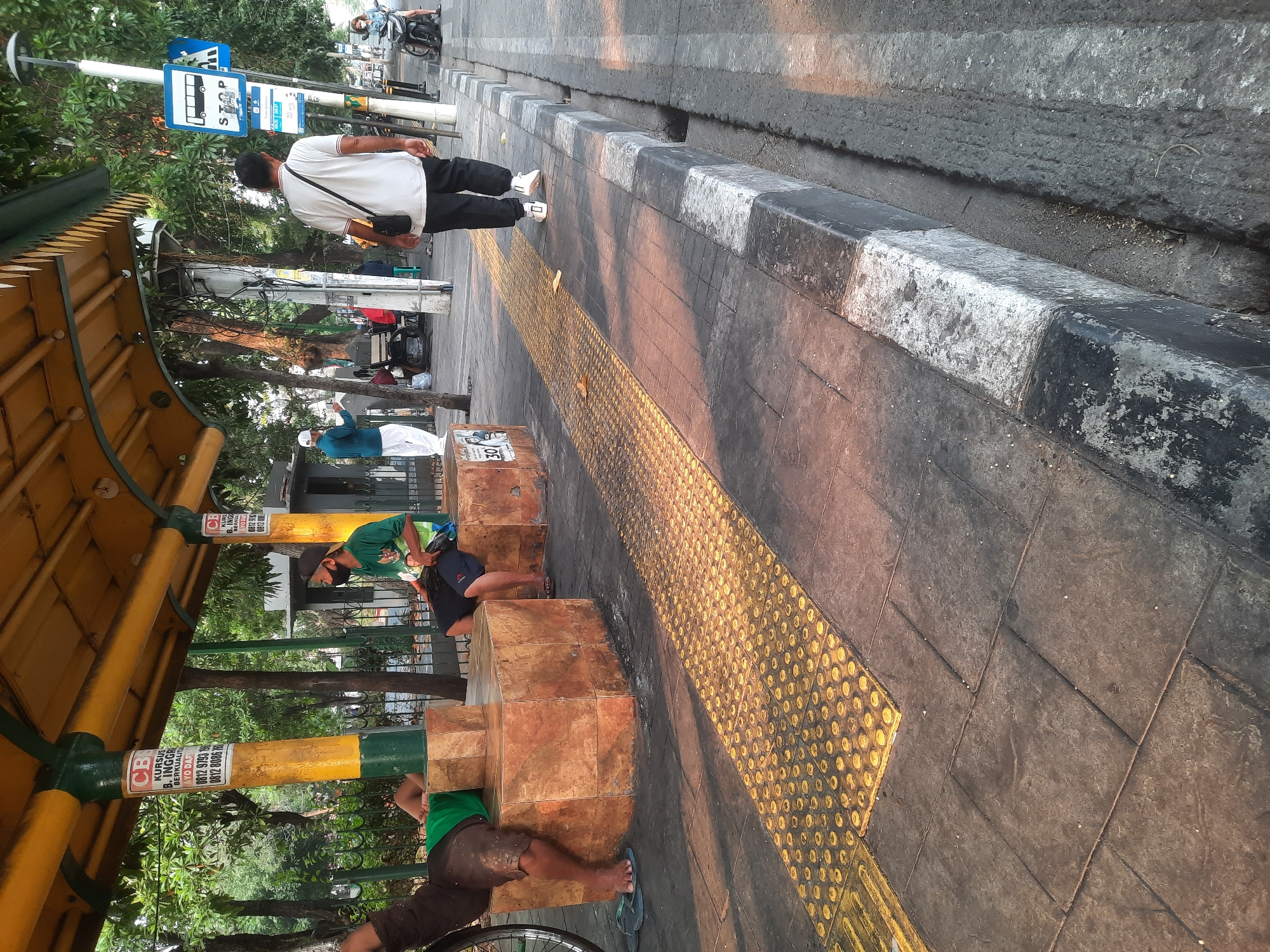 The image depicts a bus stop in North Jakarta, Indonesia, likely during daylight hours as indicated by the shadows. A covered waiting area with yellow and green supports is visible, featuring promotional posters on one of the pillars. Several individuals are present at the bus stop. One person, wearing a green t-shirt and black shorts, is seated on a textured, block-like bench and appears to be wearing a black face mask and a baseball cap. To the left, another person, clad in a green t-shirt and dark pants, is lying down on a similar bench, with their left leg extended and visible. A man in a white t-shirt and black pants, carrying a dark bag over his shoulder, walks away from the camera along the sidewalk. Further down the street, a woman on a motorcycle is seen, also wearing a face mask. The sidewalk is marked with a yellow tactile paving strip, common for visually impaired individuals. The surrounding environment includes trees and other urban infrastructure.