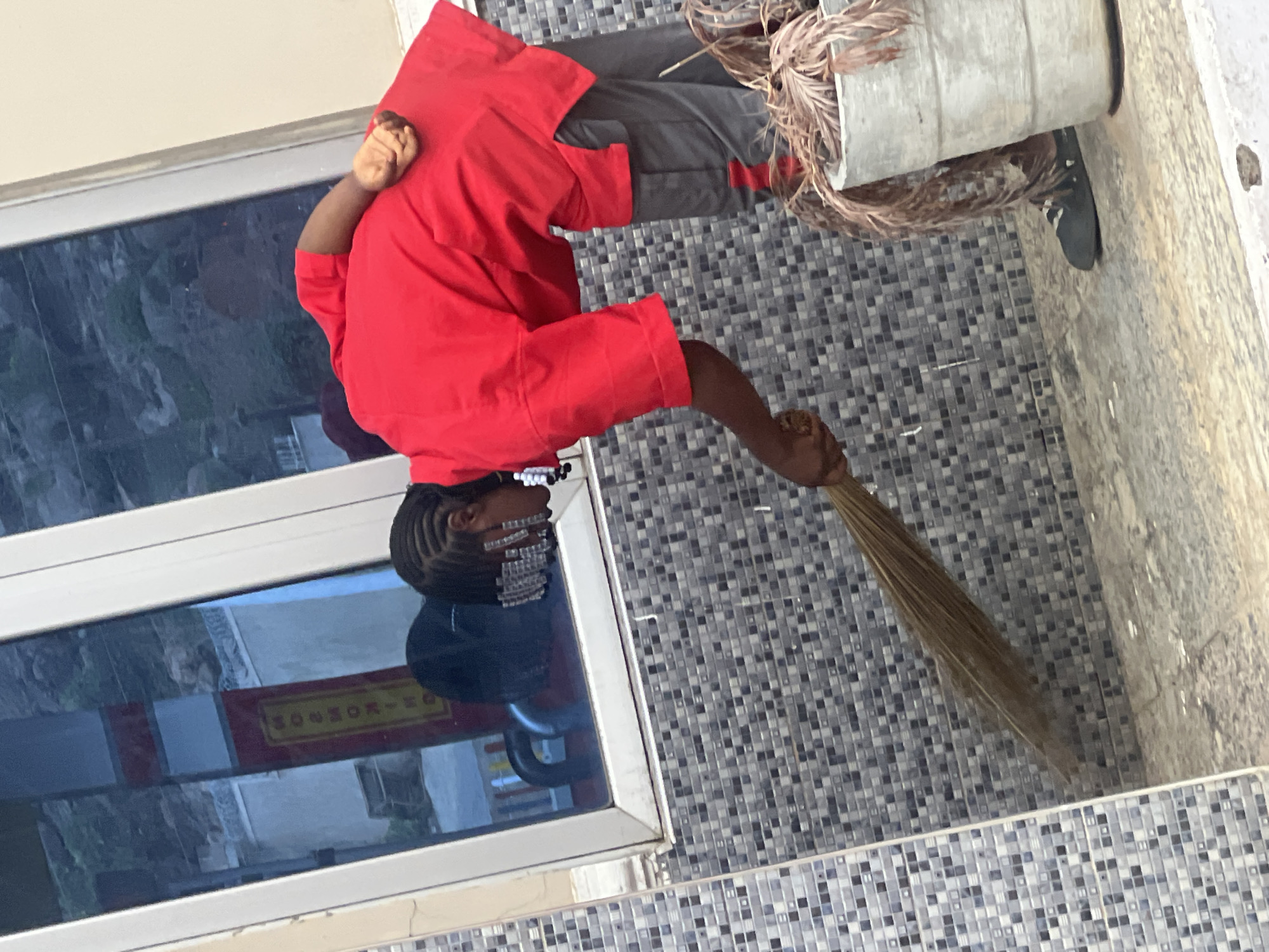 A person in a red shirt and gray pants is sweeping the floor near a window with a broom. A bucket with dried leaves is nearby. The location is possibly Gura Top, Nigeria, though this can't be confirmed definitively from the image. The floor is covered in blue and white tiles.