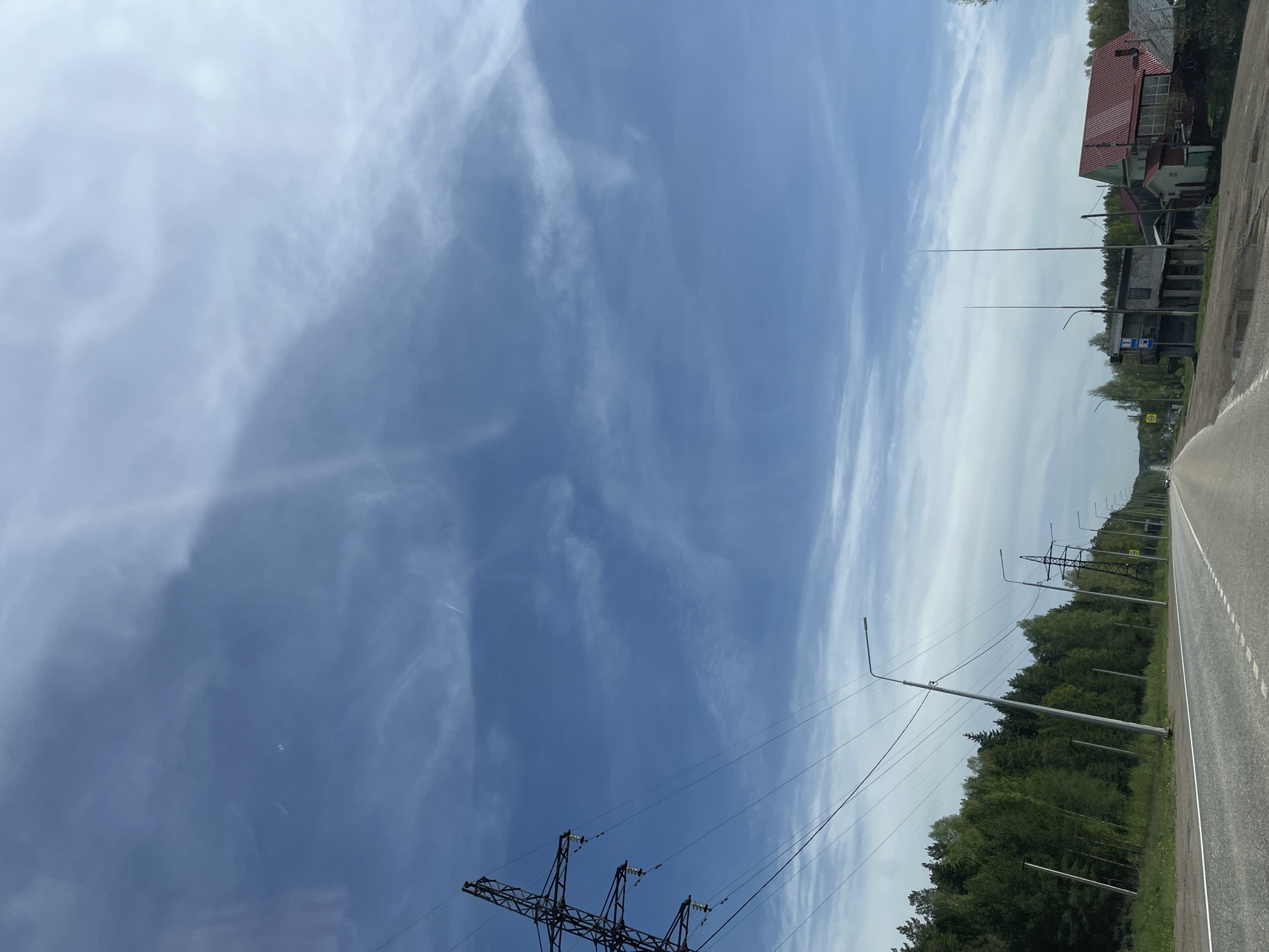 The image depicts a roadside scene in the city of Chaikovsky, Russia. A two-lane asphalt road runs along the right side of the frame, marked with a white line. Adjacent to the road is a strip of green vegetation, composed of a mix of trees and bushes. Above the trees, a series of utility poles with electrical lines are visible, stretching into the distance. On the left, a large metal electrical transmission tower is partly visible. The sky dominates the upper portion of the image, displaying a pattern of blue patches interspersed with streaky white clouds. In the upper right corner, a section of a red-roofed building and other structures indicate a nearby populated area, including a bus stop sign.