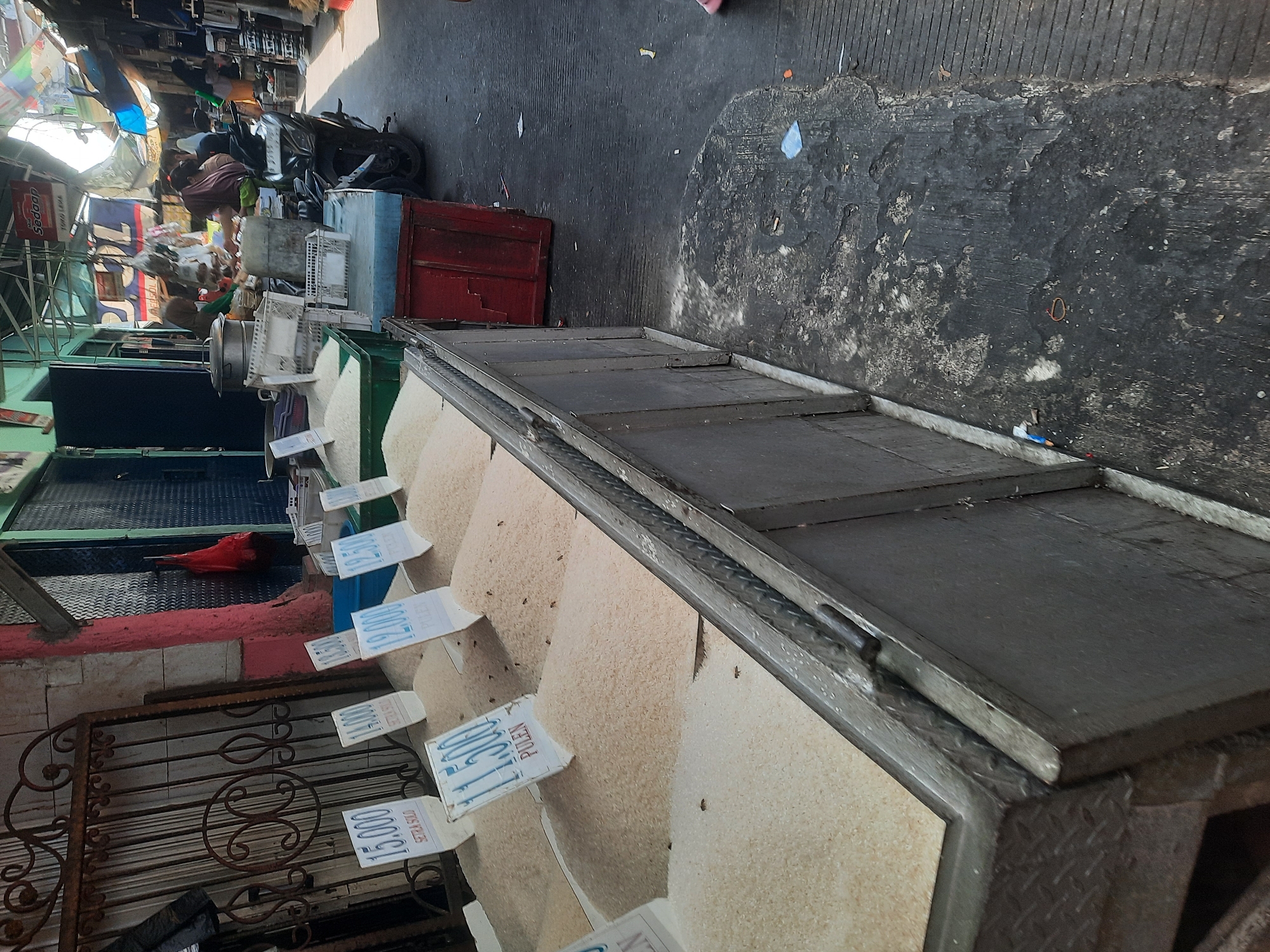 The image depicts a street-level market stall in North Jakarta, Indonesia. The stall features several large piles of rice, each accompanied by a sign indicating the price. The signs show prices such as "15.000," "14.000," "12.000," and "11.500," with the word "PULEN" or "SETRA SOLO" appearing below the numbers, likely indicating rice varieties or quality. The rice is displayed in shallow, open containers or directly on the ground, covered with protective sheeting in some instances. In the background, a motorcycle is parked, and several people are visible tending to stalls or interacting. The environment appears to be an outdoor market with tarpaulins and awnings providing shade. The ground is paved with concrete, showing signs of wear and debris. The overall scene suggests a bustling local marketplace.