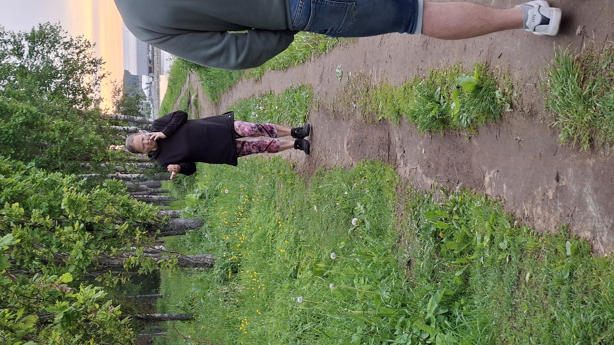 A woman in a black hooded sweatshirt and patterned leggings is walking on a dirt path and talking on a mobile phone. She is gesturing with her right hand. She is surrounded by green grass and trees on the left side of the path, and a body of water with a large structure in the background on the right. The sky in the background is a pale orange, suggesting it is sunset or sunrise. Another person's legs and shorts are visible in the foreground on the right side of the image. The scene appears to be in an outdoor recreational area, possibly a park or a path along a waterfront.