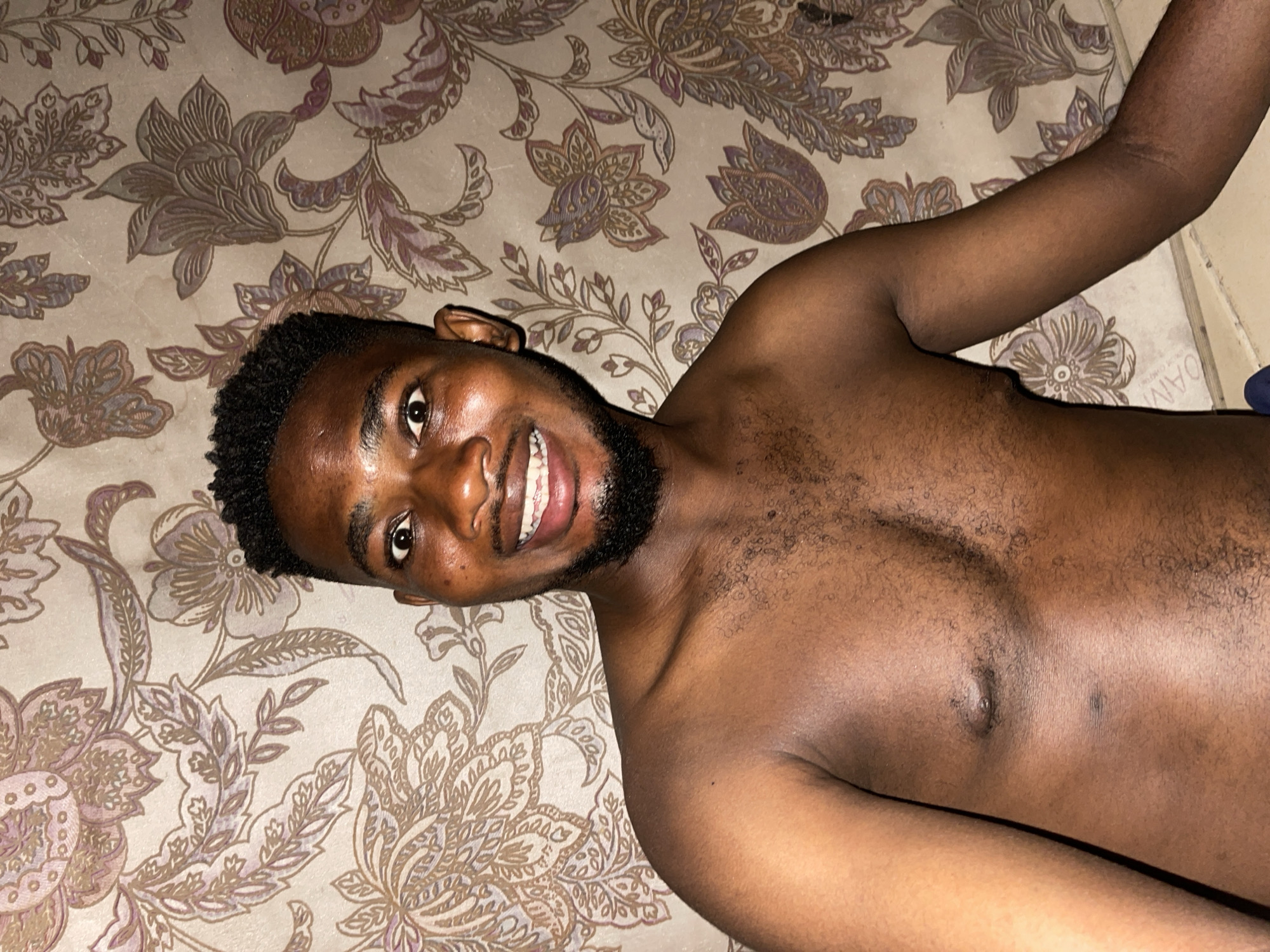 A shirtless man is lying on a patterned surface, possibly a bed or couch, indoors. He is dark-skinned with short, dark hair and a beard, and he is smiling. The background is a floral pattern in shades of brown, beige, and purple. The location is possibly Unguwar Gabas, Nigeria.